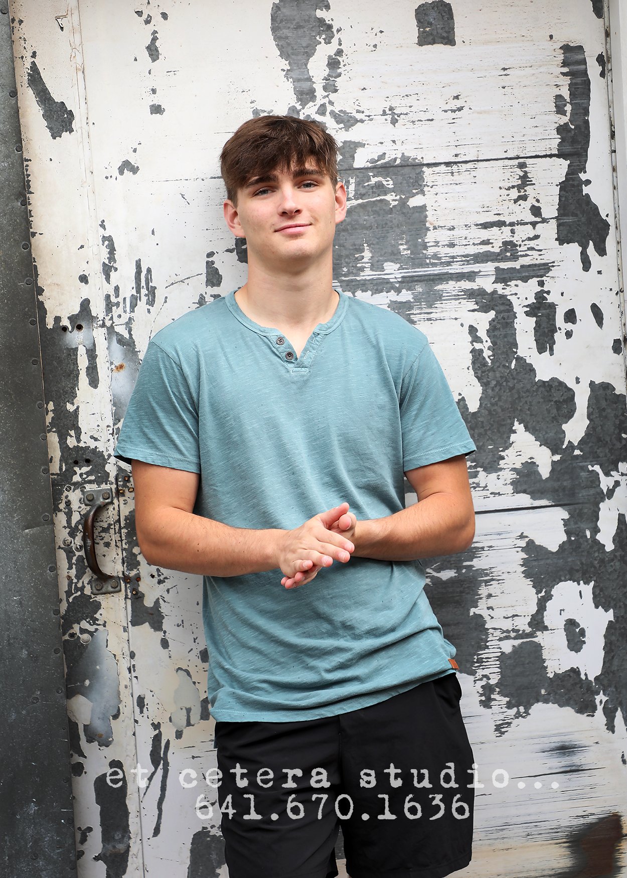 A young man with brown hair wearing a blue T-shirt and black shorts standing in front of a weathered metal wall with peeling paint, with his hands clasped together.