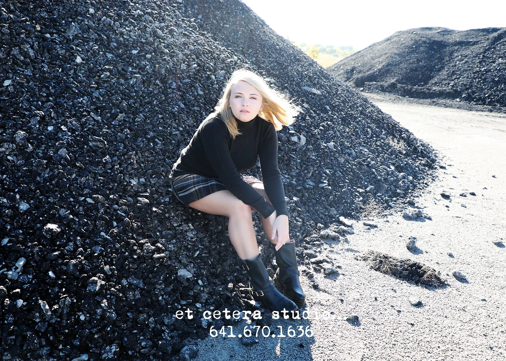A young woman with blonde hair sitting on a tall pile of black gravel or coal, wearing a black turtleneck, plaid skirt, and black boots, with a bright background and a gravel road nearby.