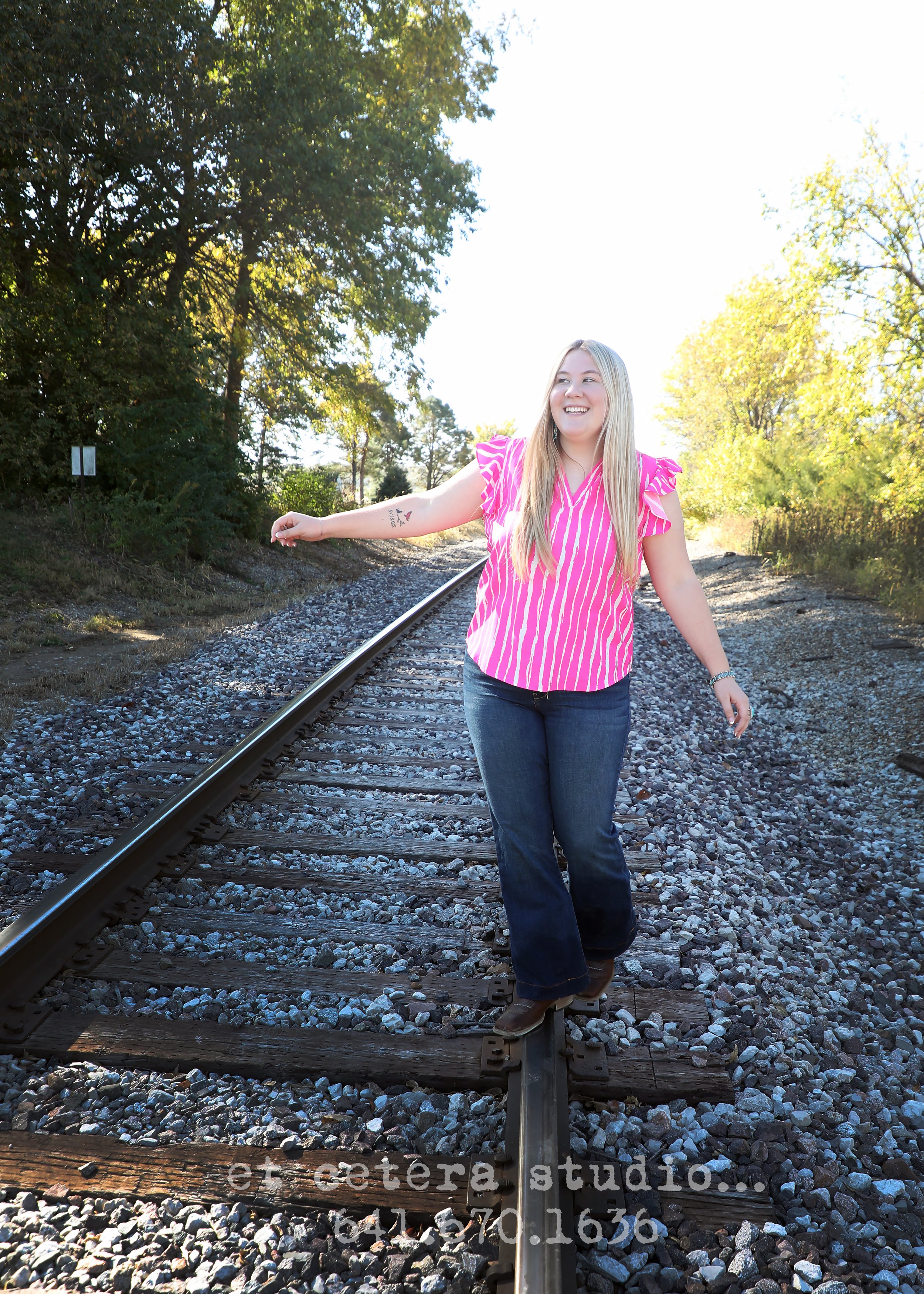 A young woman with long blonde hair posing on a railroad track, wearing a pink and white striped blouse, jeans, and brown boots, smiling and extending her left arm to the side, with trees and greenery in the background on a sunny day.
