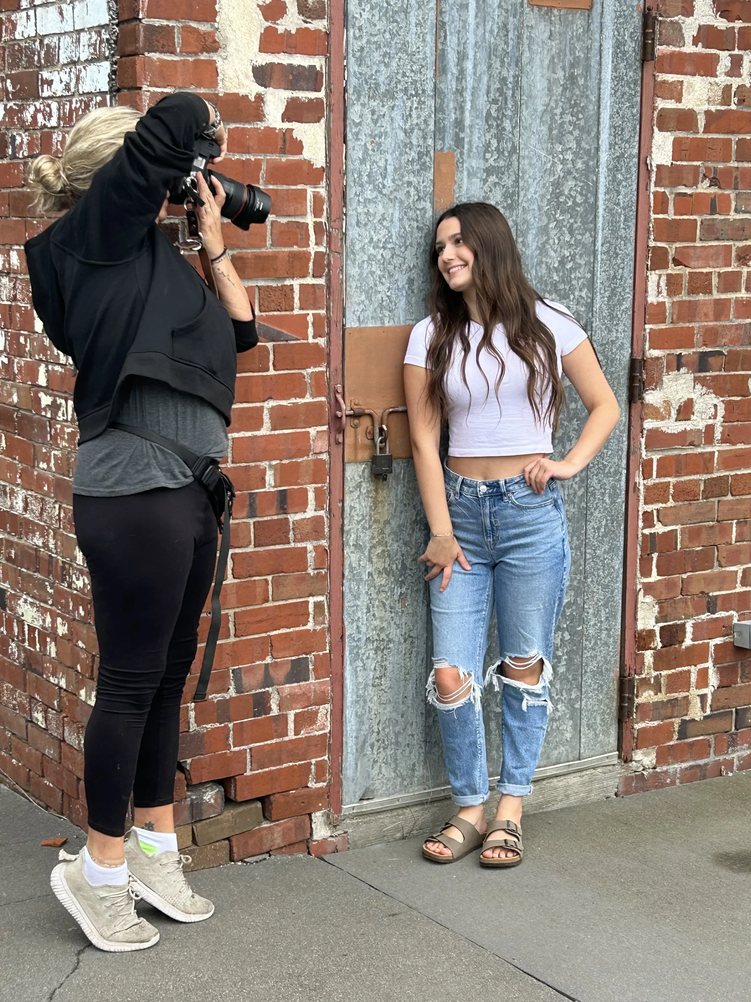 A woman with long dark hair is smiling and posing against a weathered blue door with graffiti. A photographer with blonde hair pulled back and wearing black clothing is taking her picture. The scene is set outdoors on a brick-lined wall with concrete pavement.