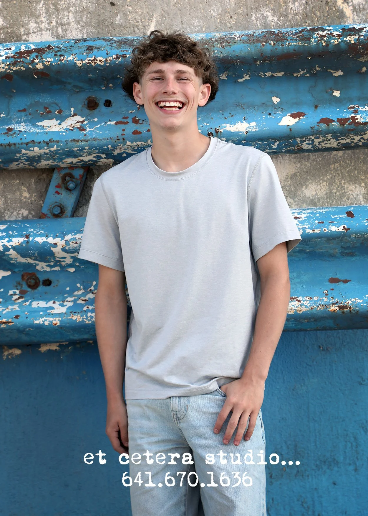 Young man with curly hair smiling and leaning against a weathered blue wall.