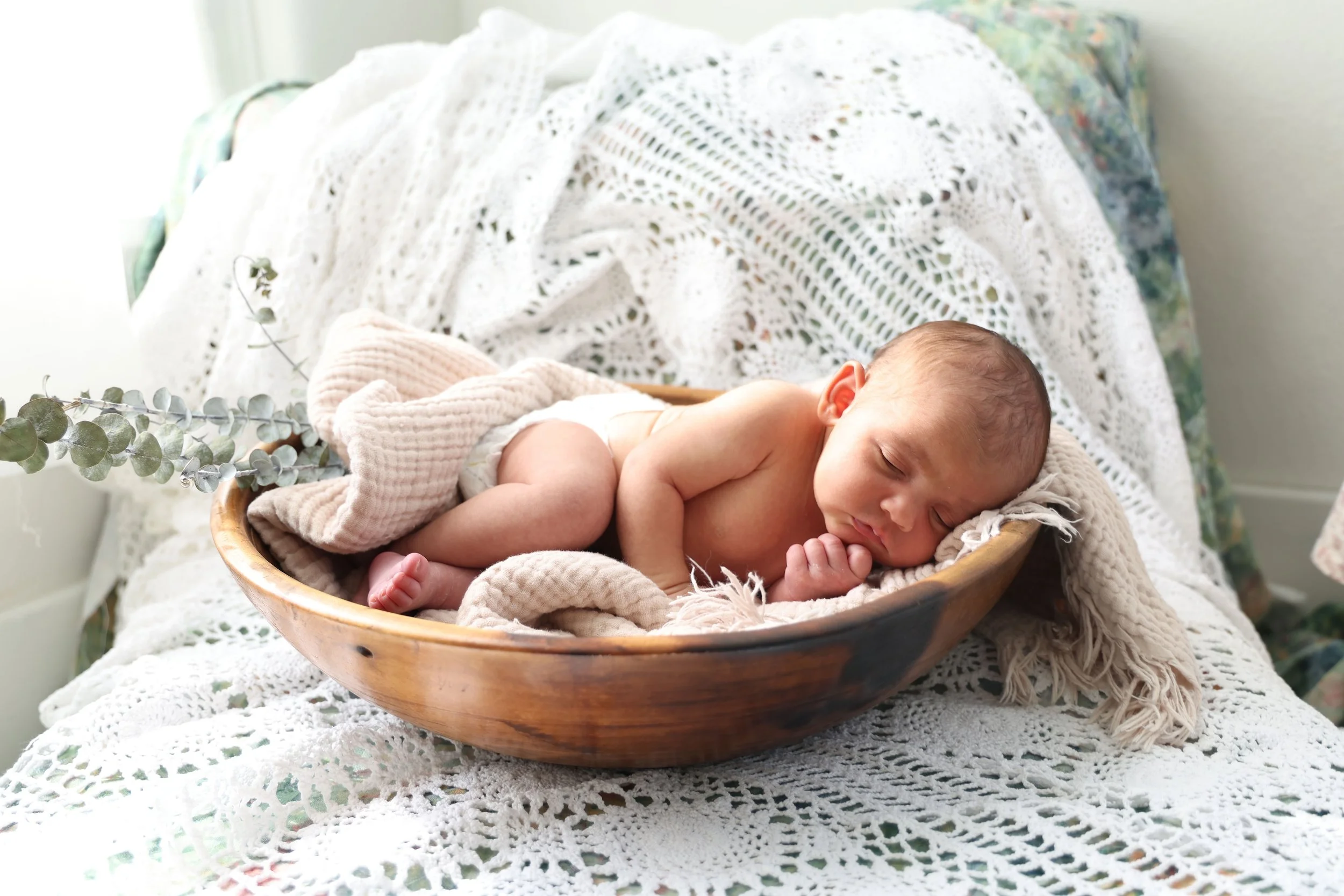 A sleeping newborn baby lying in a wooden bowl on a white crocheted blanket with green leaves in the background.