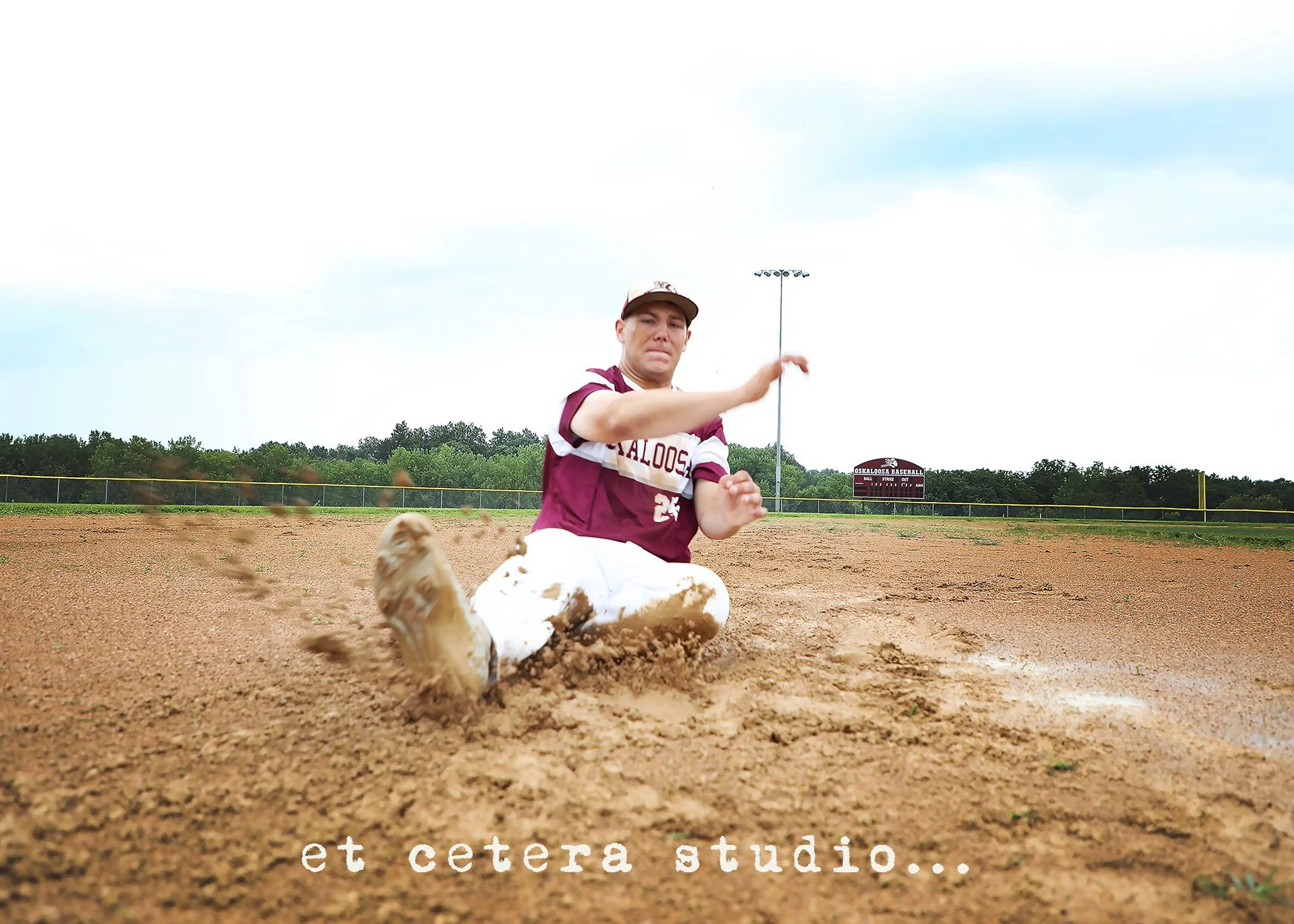 A baseball player wearing a maroon and white uniform with the number 24 is sliding into a base on a dirt field, with a cloudy sky above and a scoreboard in the background.