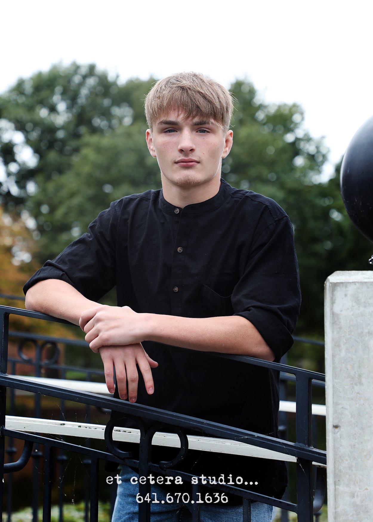 A young man with light brown hair and fair skin, wearing a black button-up shirt, leaning on a black metal fence outdoors with trees in the background.