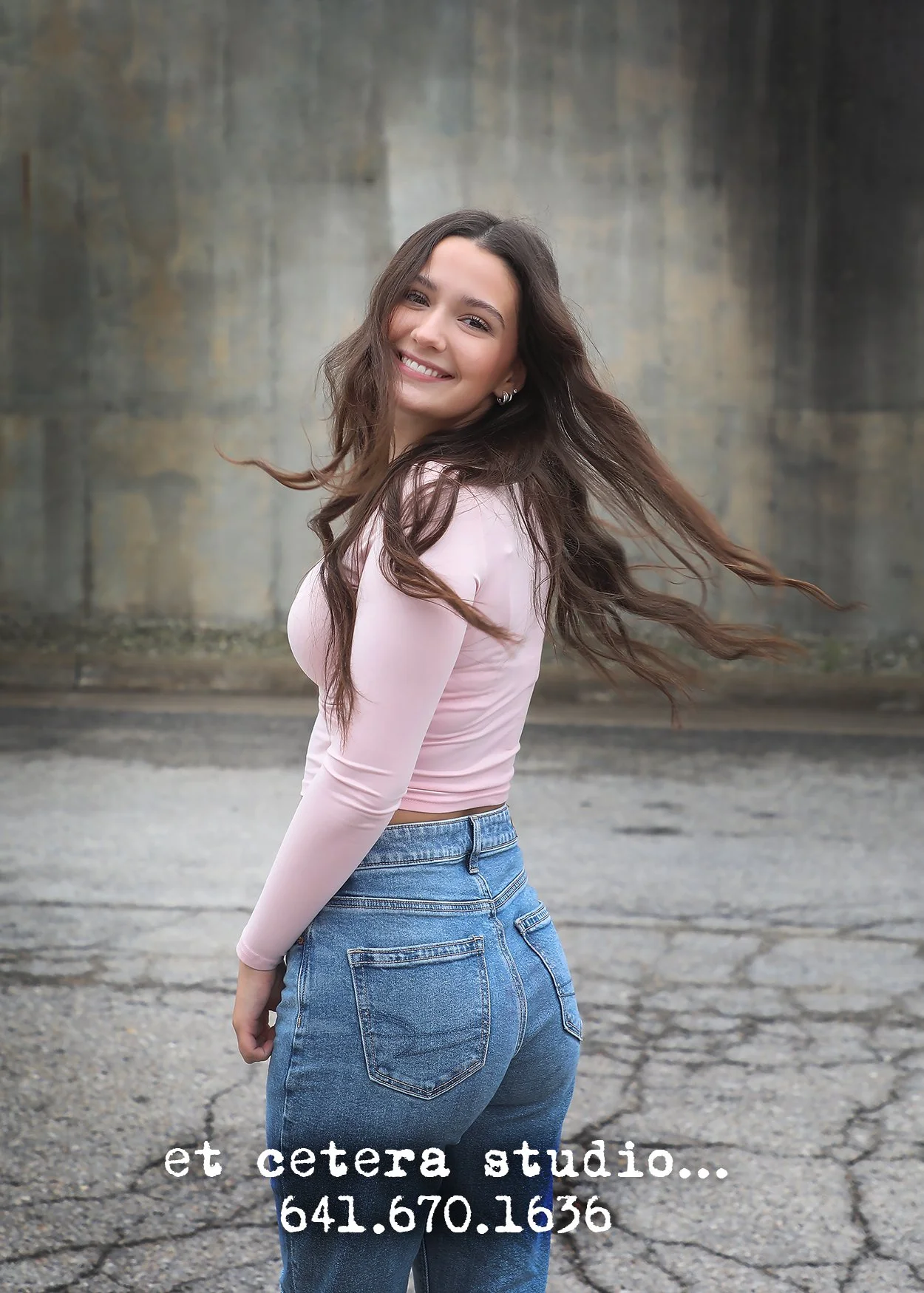 Young woman with long brown hair smiling outdoors in front of a concrete wall, wearing a pink long-sleeve top and blue jeans.