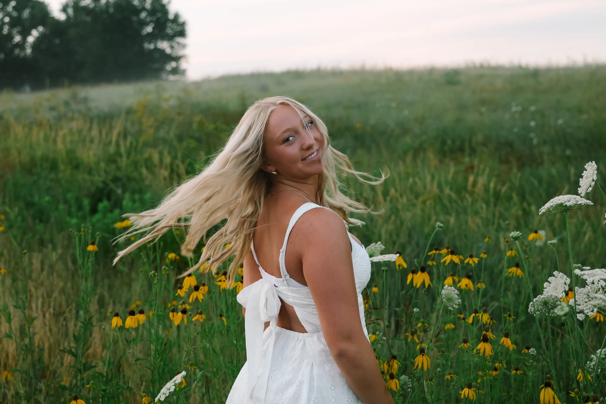 A smiling woman with long blonde hair wearing a white dress standing in a field of yellow and white wildflowers with trees and a cloudy sky in the background.