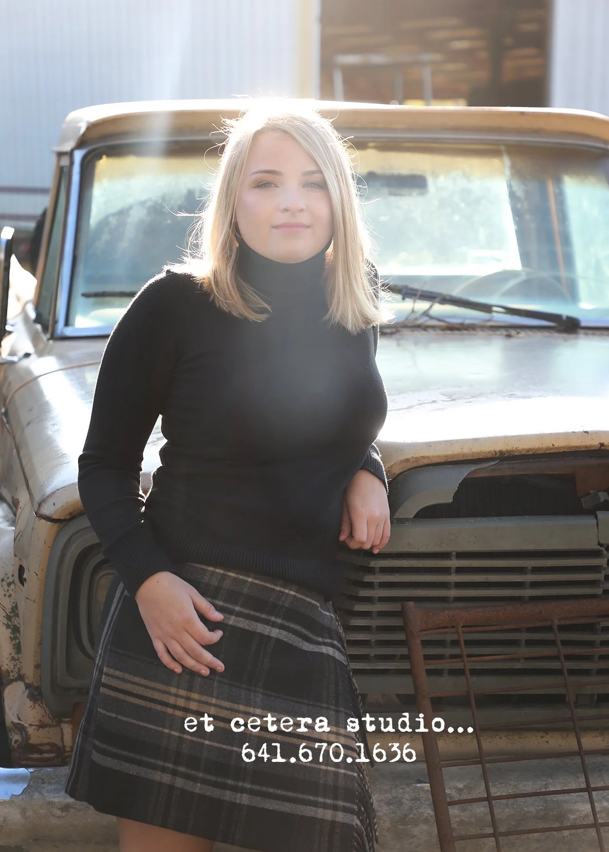 A young woman with blonde hair, wearing a black turtleneck sweater and a plaid skirt, stands in front of an old, rusty vehicle, possibly a truck, in an outdoor setting with a barn-like structure in the background. Text overlay reads 'et cetera studio