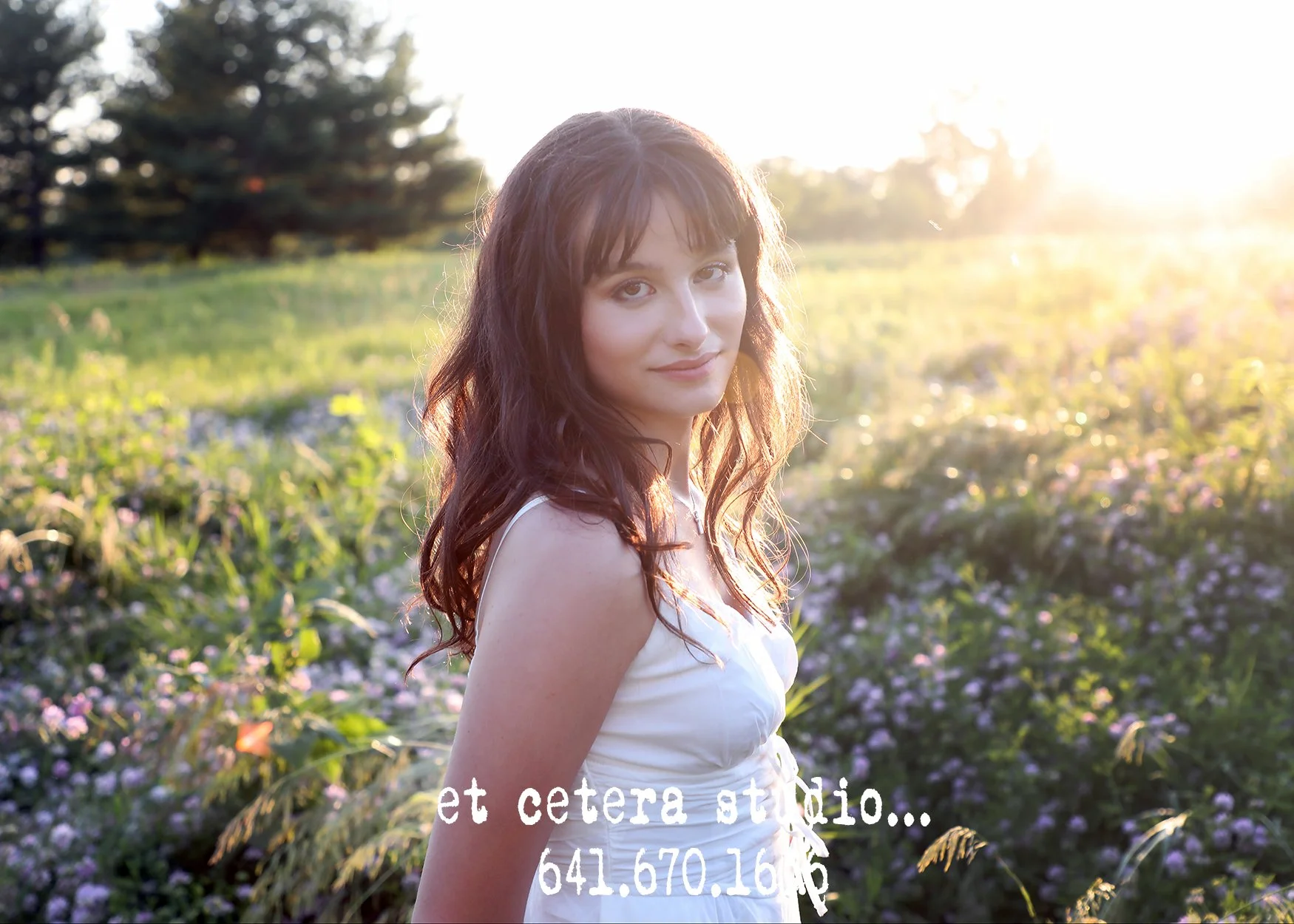 A young woman with shoulder-length dark hair and light skin stands in a field of purple flowers during sunset, wearing a white sleeveless dress, facing slightly to the right with a gentle smile.