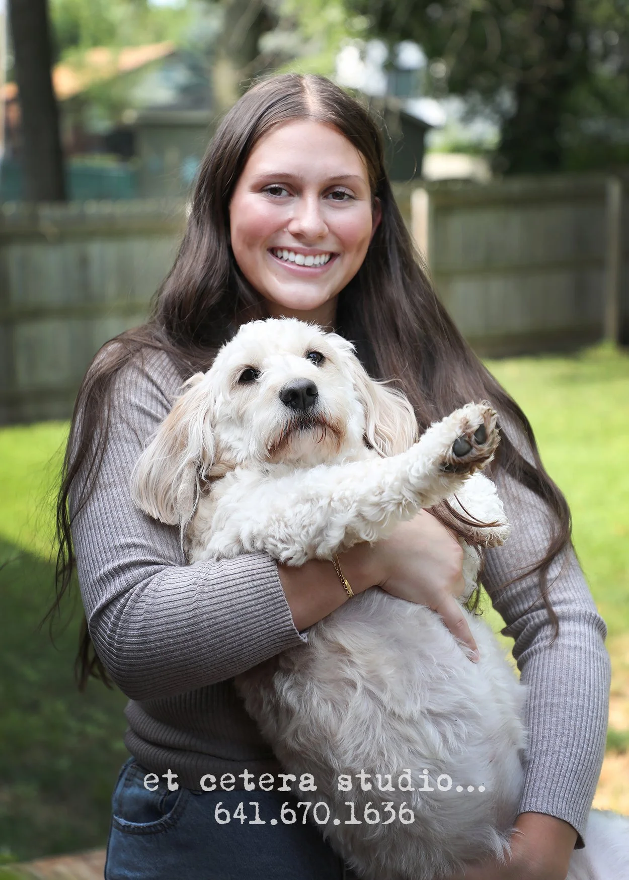 A young woman with long brown hair smiling while holding a fluffy white dog outdoors in a yard with a wooden fence and trees in the background.