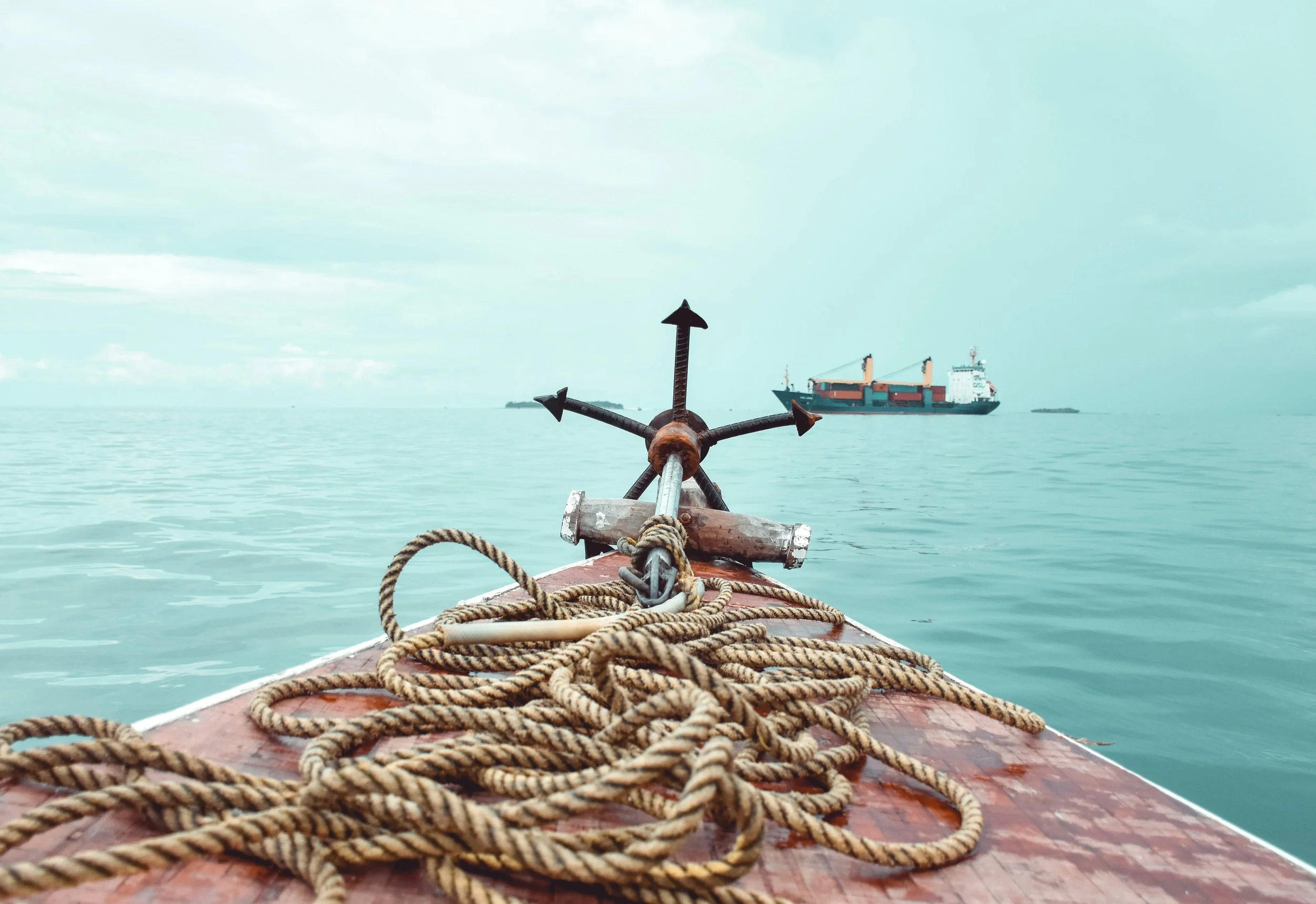 View from the bow of a boat with ropes, looking out onto open water with a large cargo ship in the distance and an island ahead.