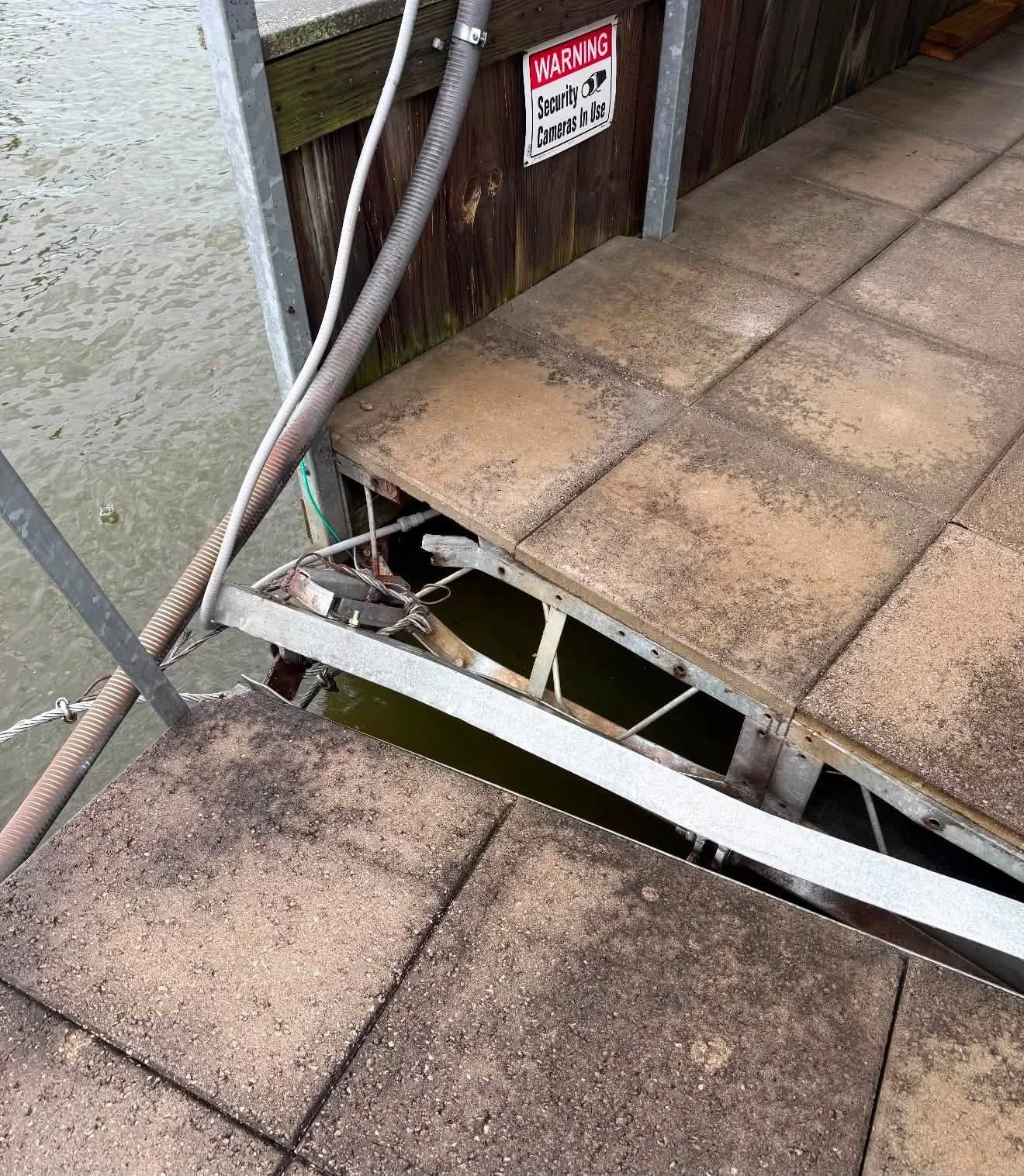 Weathered dock and walkway with exposed metal framework and air or water pipes. Sign on wooden wall warns of security cameras in use.
