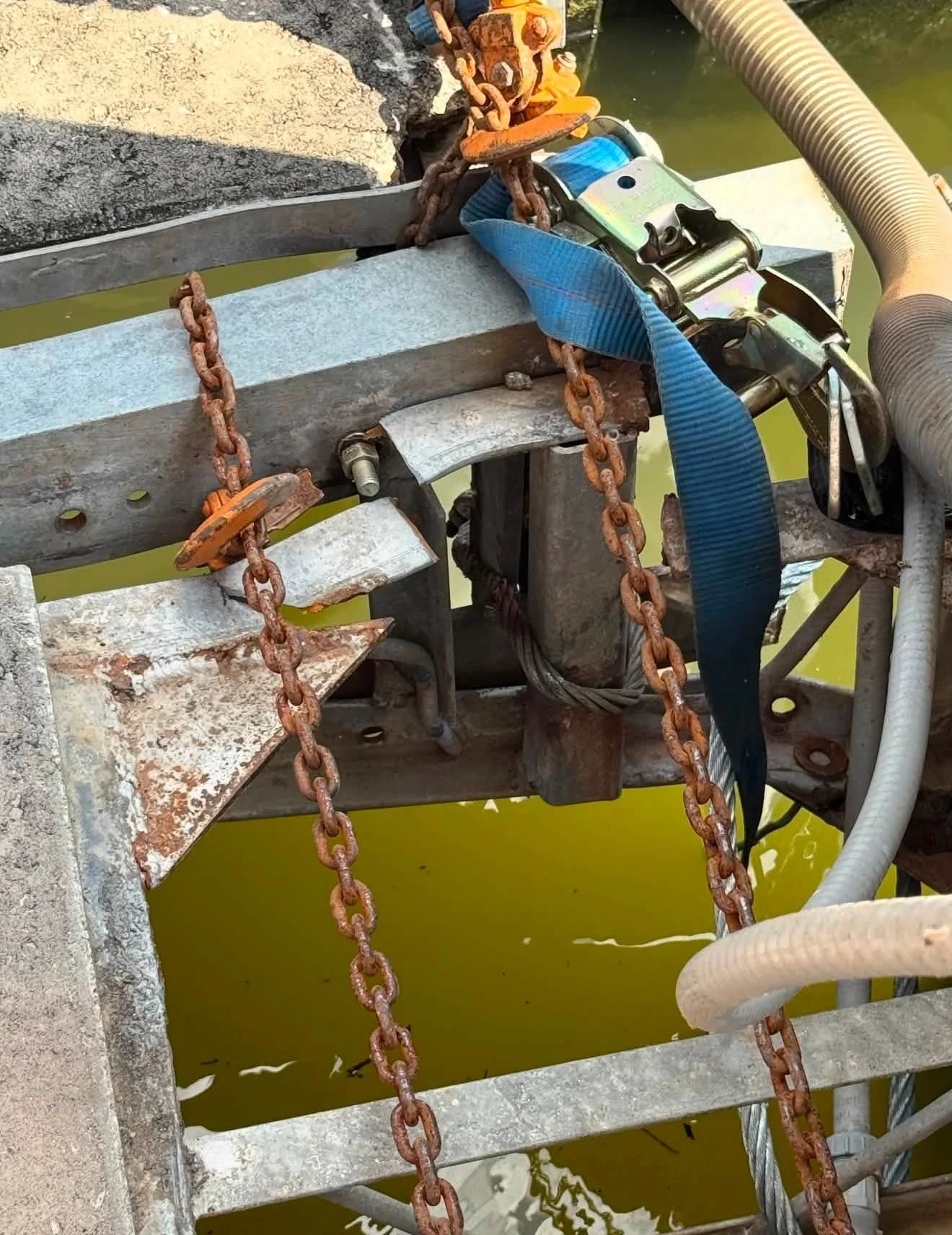 Close-up of a rusty metal structure with chains, pipes, and mechanical parts, over water with a greenish tint.