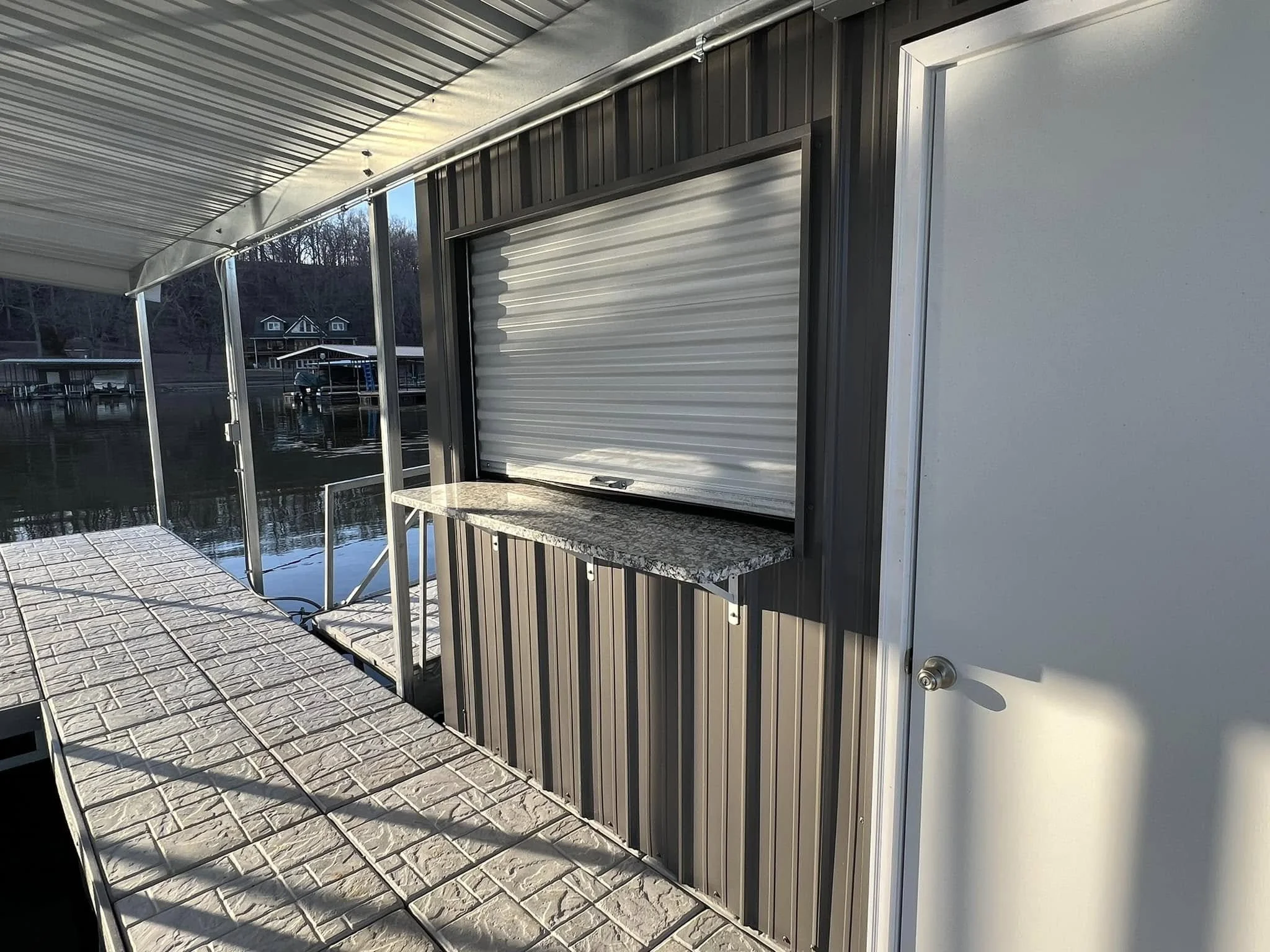 View of a boat docked at a canal with houses and trees in the background, a covered patio with patterned concrete tiles, and a window with a metal rolling shutter on a building.