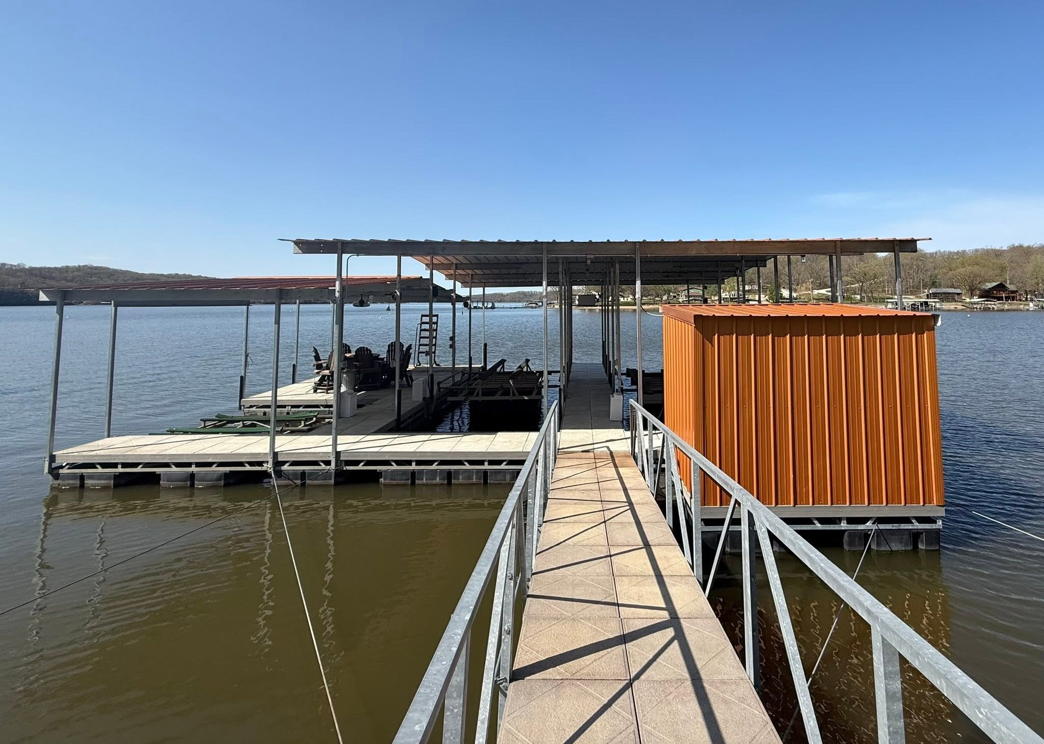 View of a dock with a covered boat slip extending over a lake, featuring a small orange shed, with houses and trees on the opposite shore under a clear blue sky.