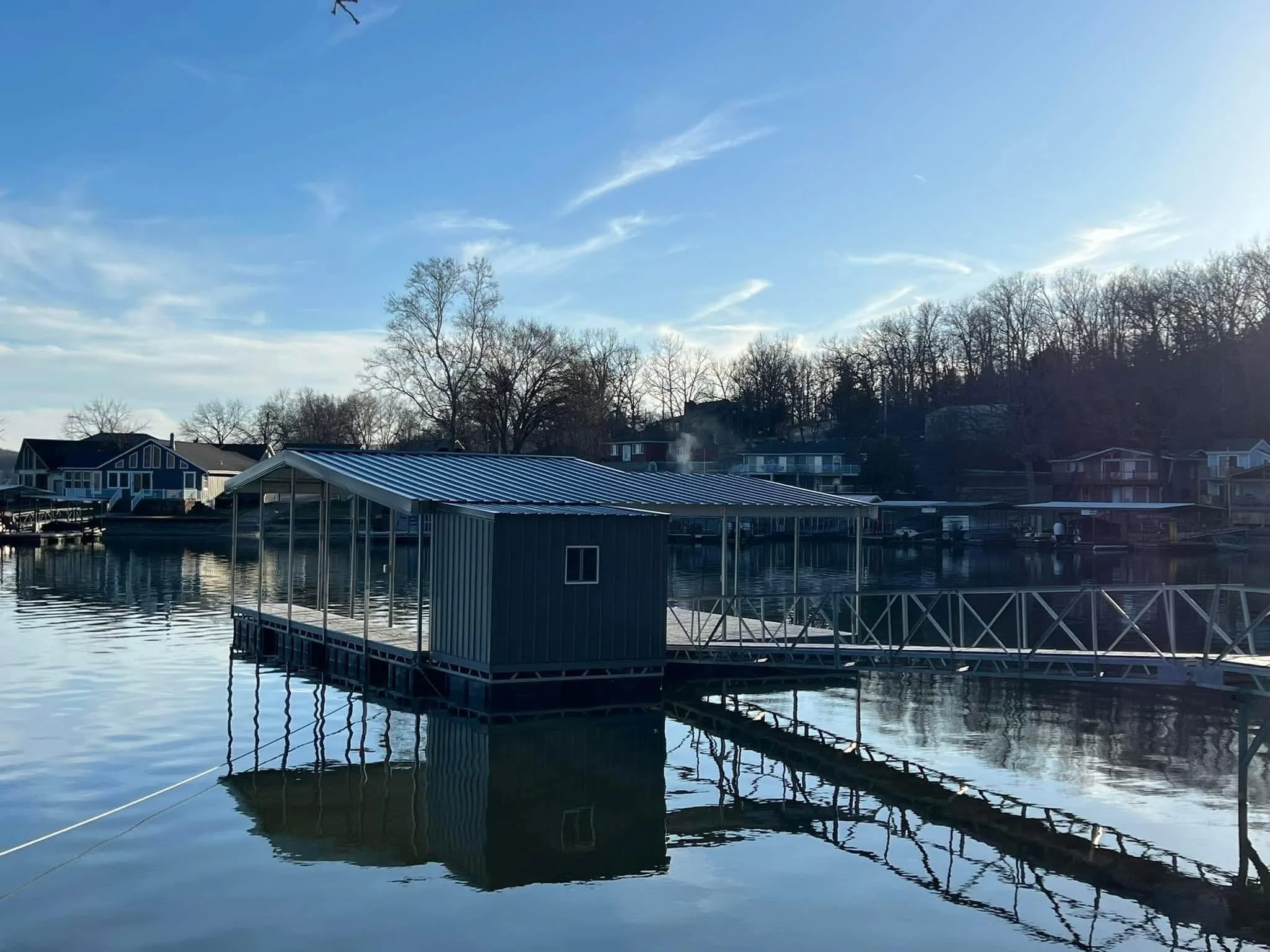Calm water reflects a floating dock and a waterfront house with a blue roof, situated among trees with bare branches, under a clear blue sky.