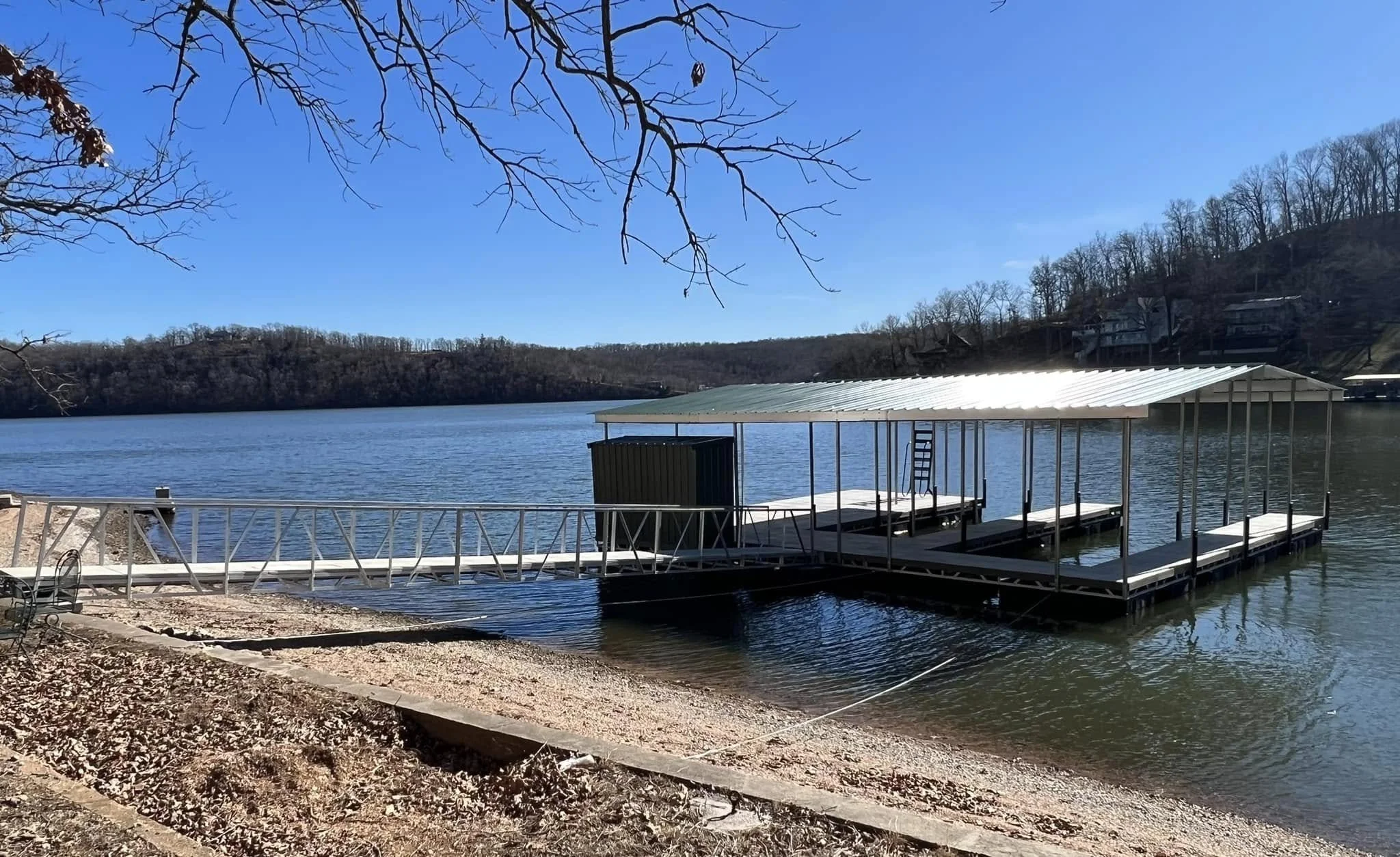 A lakeside scene with a floating dock and a boat shelter, bare trees in the background on a clear, sunny day.