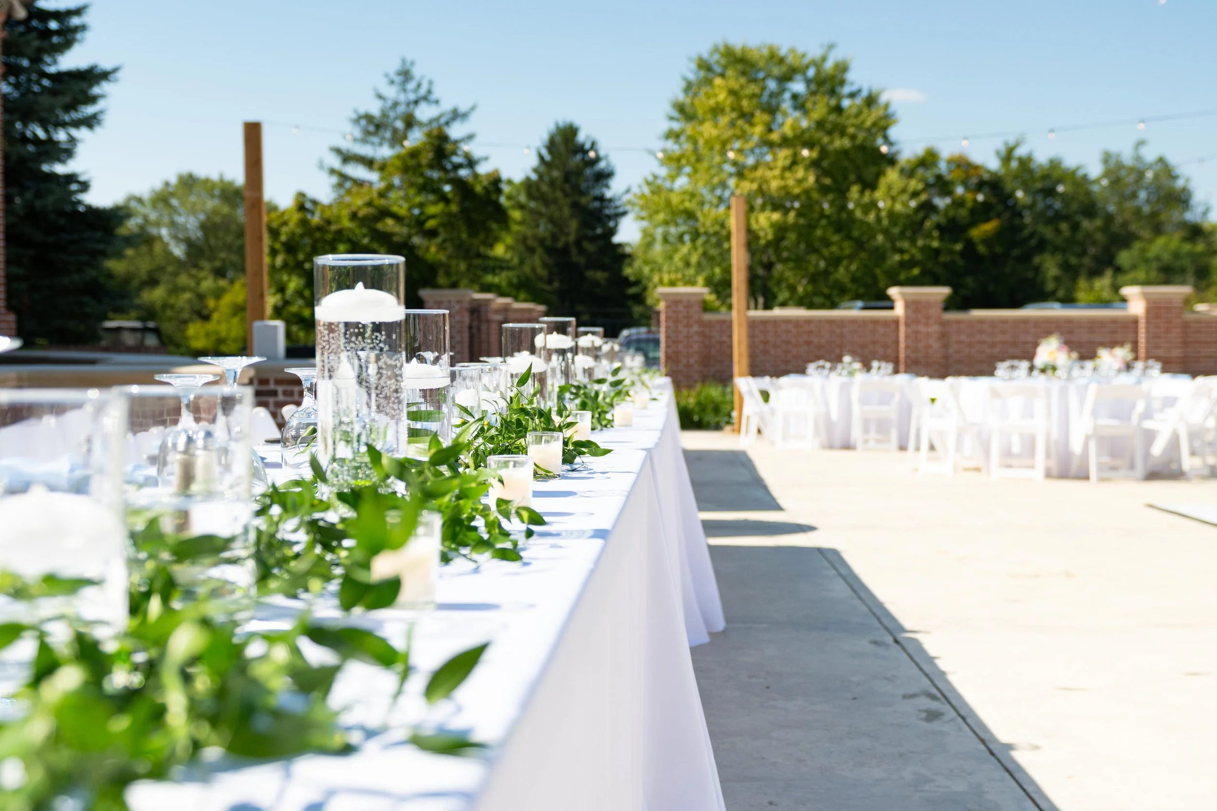 Outdoor banquet setup with a long white table adorned with green foliage and glass candle holders, set against a backdrop of trees, brick wall, and clear blue sky.