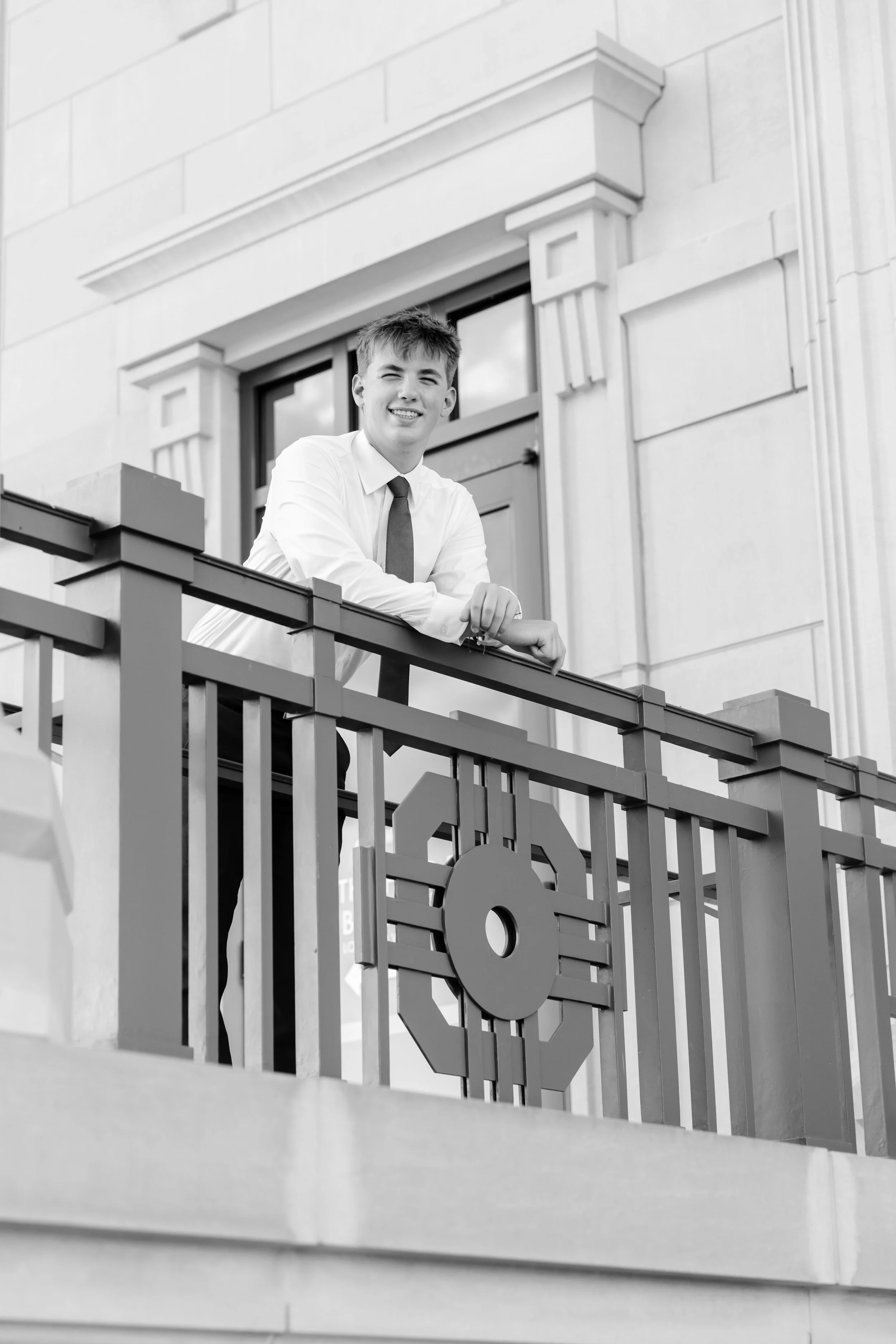 A young man in a white shirt and tie leaning on a decorative railing outside a building with a large window and ornate architectural details.