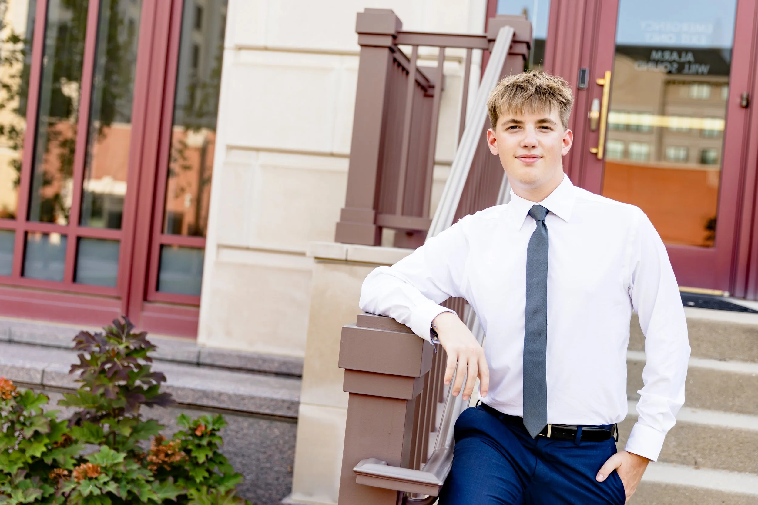 A young man in a white shirt and gray tie leaning against a brown railing outside a building with red trim and large windows. There are steps leading up to the entrance and some plants in the foreground.