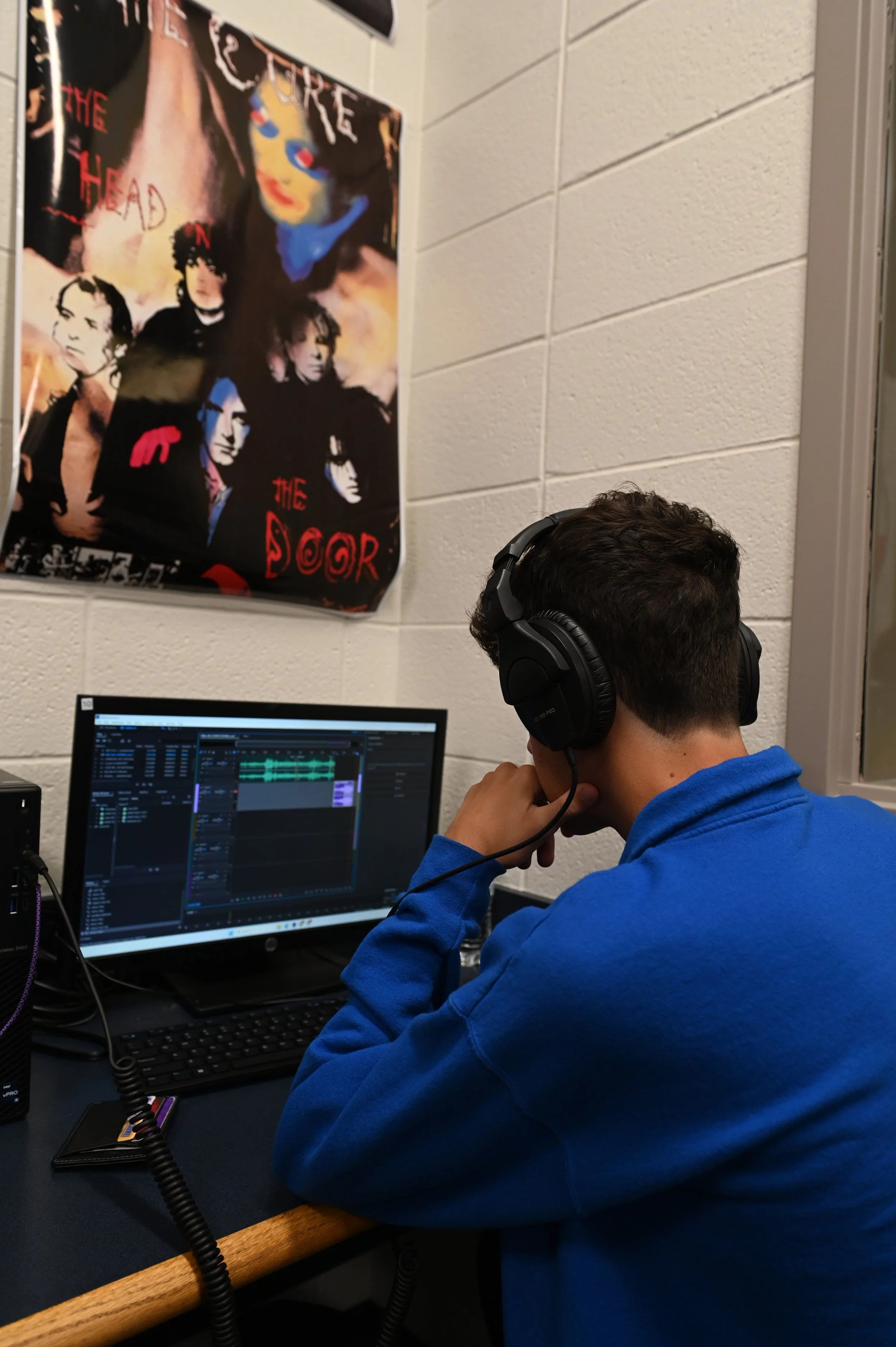 A person wearing a blue hoodie and large over-ear headphones working on a music editing software on a computer in a room with white brick walls and a colorful poster on the wall.