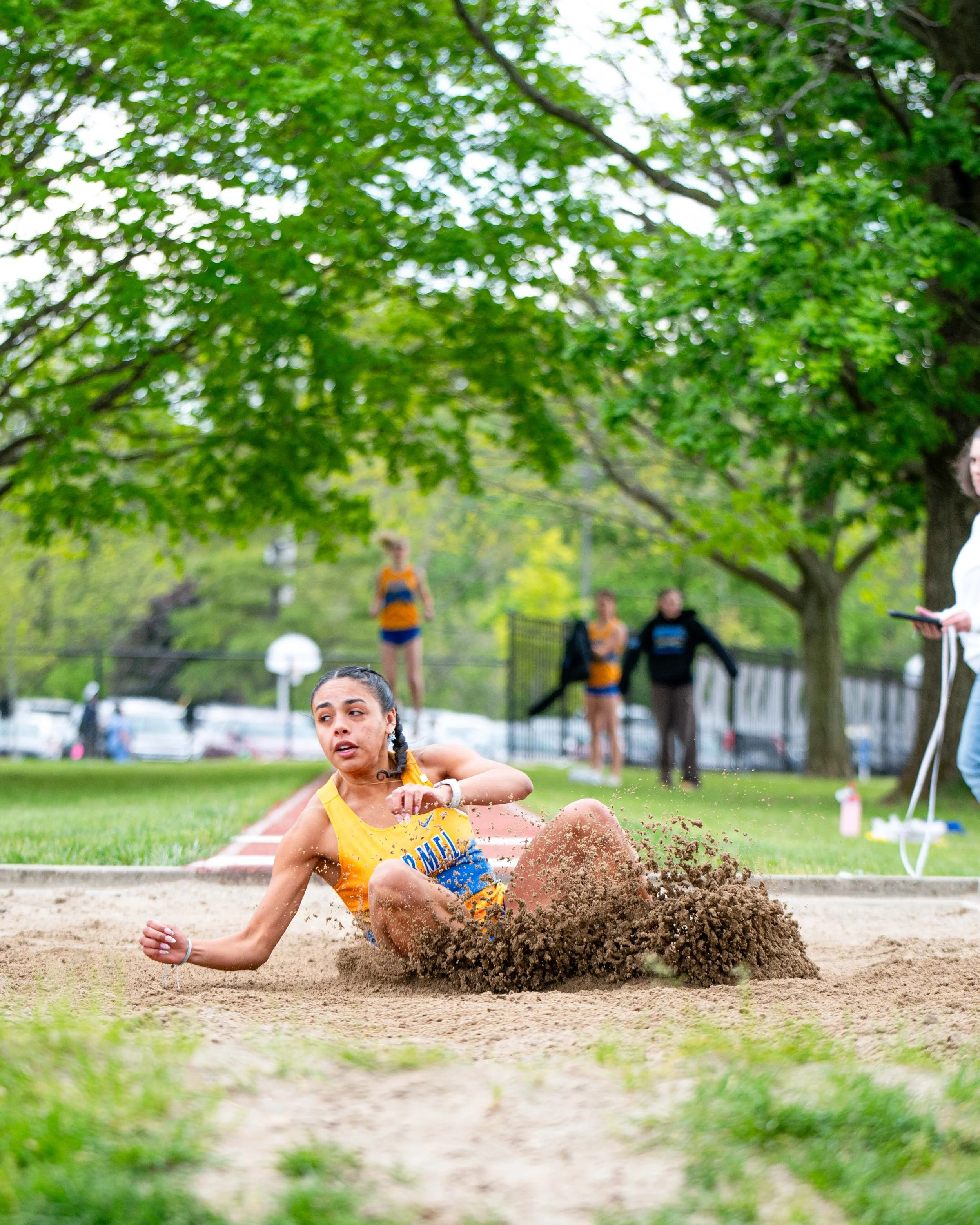 Male long jumper in orange and blue uniform landing in sandpit during outdoor track event, with trees and spectators in background.