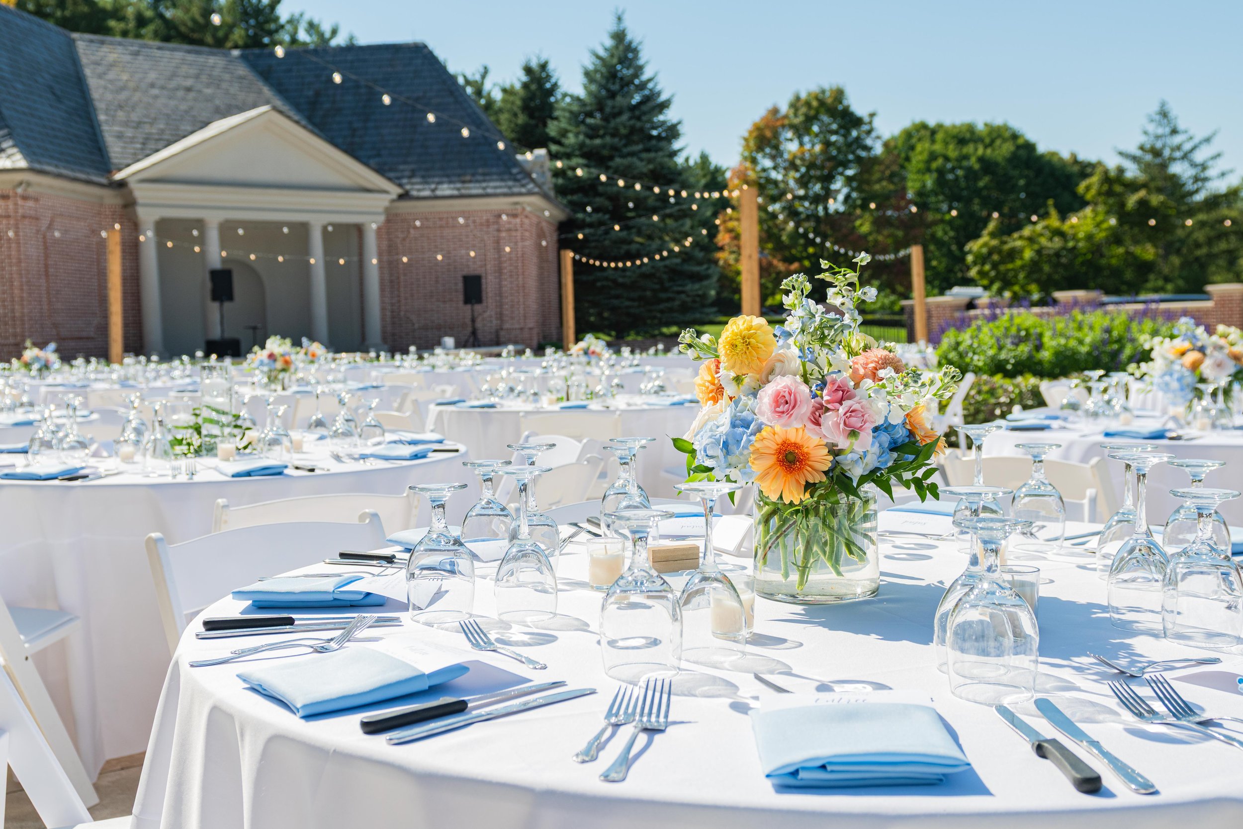 Outdoor wedding reception setup with round tables draped in white tablecloths, each adorned with a floral centerpiece, glassware, and neatly folded napkins. String lights hang above, and a brick building with a porch is in the background, surrounded 