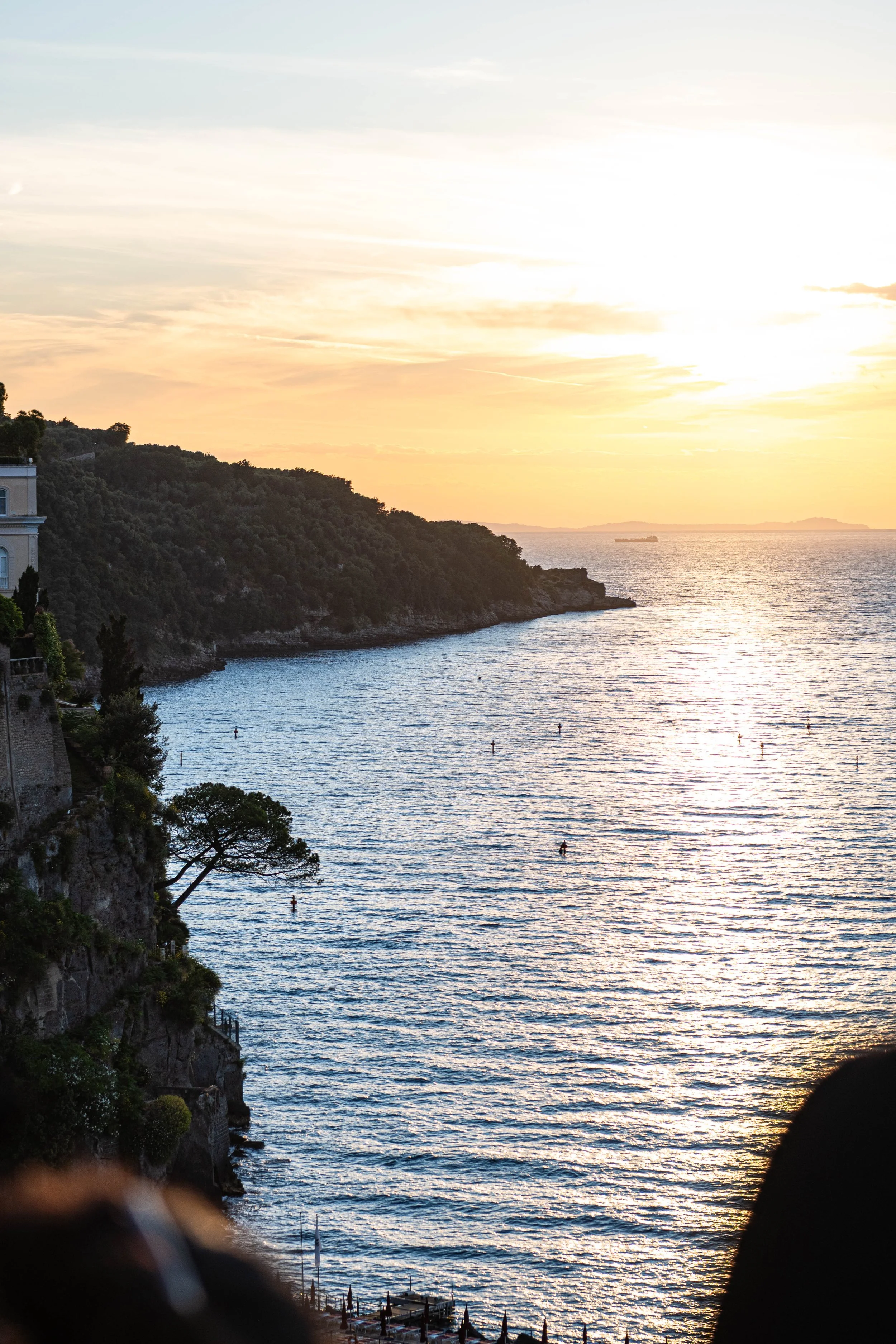 Sunset over a body of water with a ridged coastline and scenic trees. A building is visible on the left at the cliff's edge, and there are some structures and umbrellas near the water's edge.