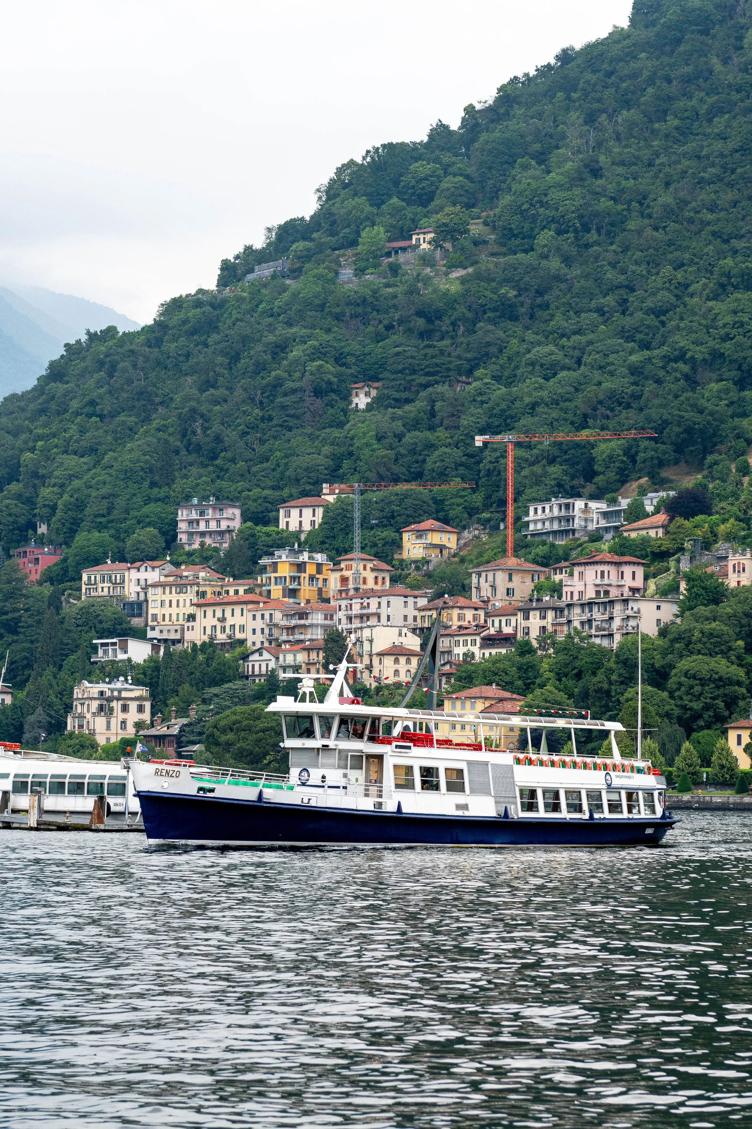 A boat named Renzo sailing on a lake with houses and a wooded hillside in the background.