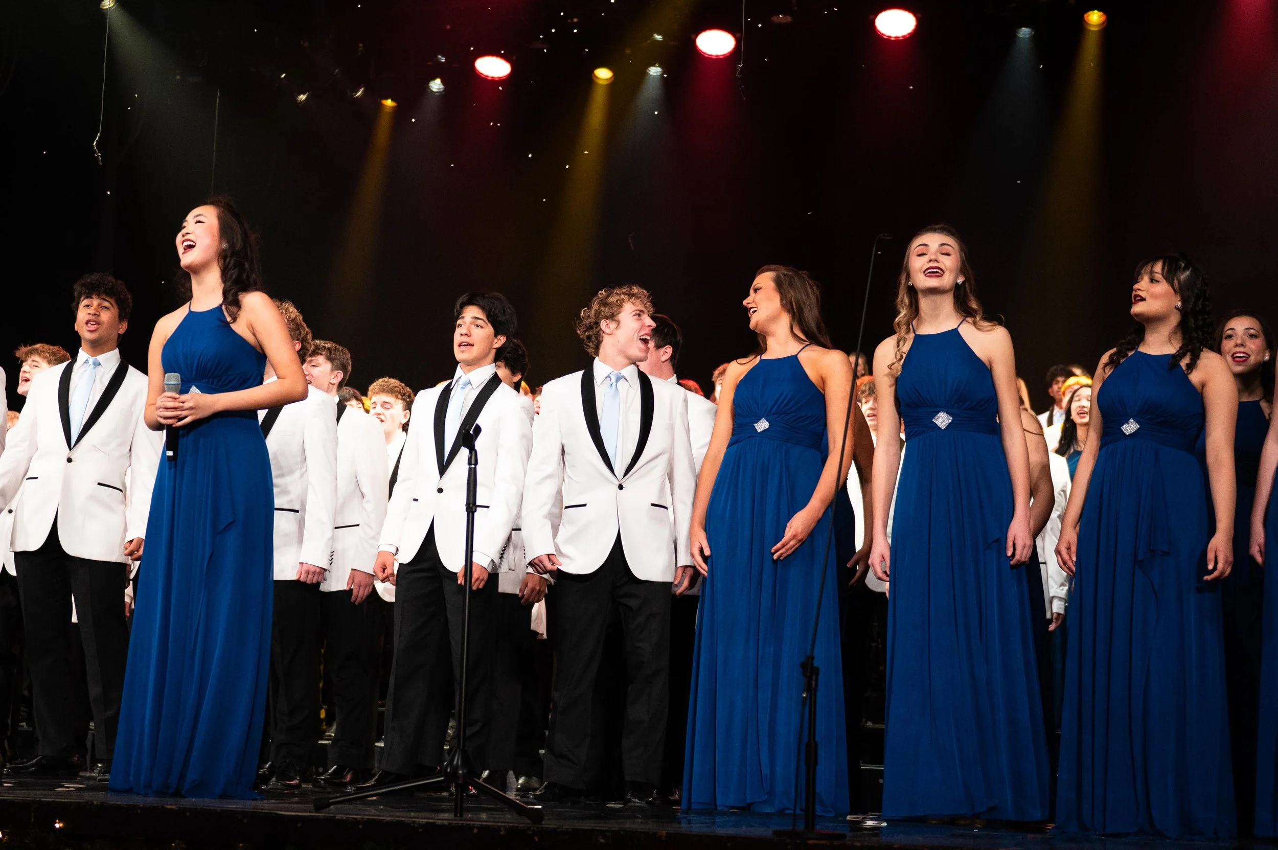 Group of young performers on stage, smiling and singing, with girls in blue dresses and boys in white suits and black ties, under stage lights.