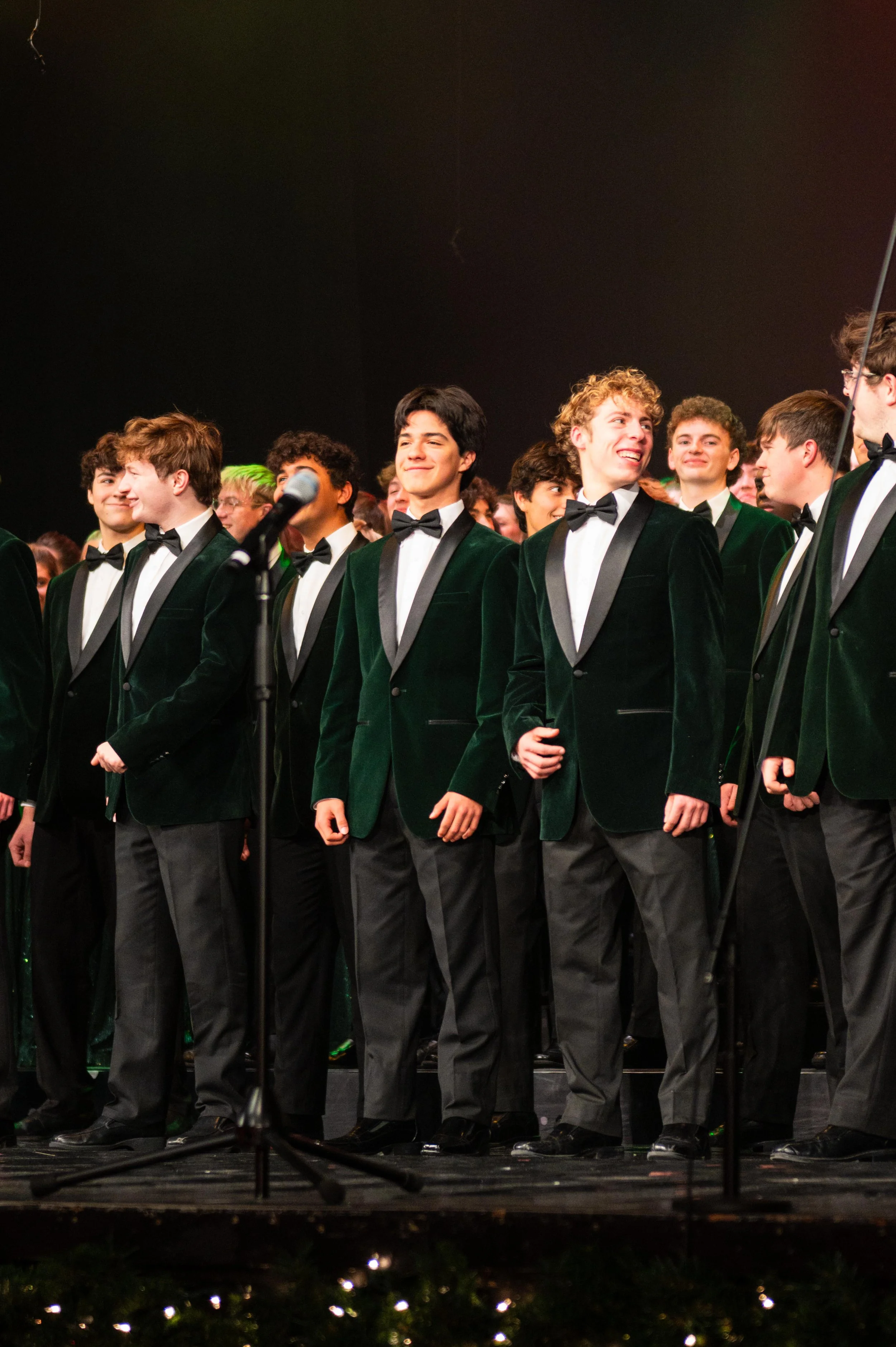 A group of young men dressed in tuxedos with black bow ties, standing on stage during a choir or orchestra concert.
