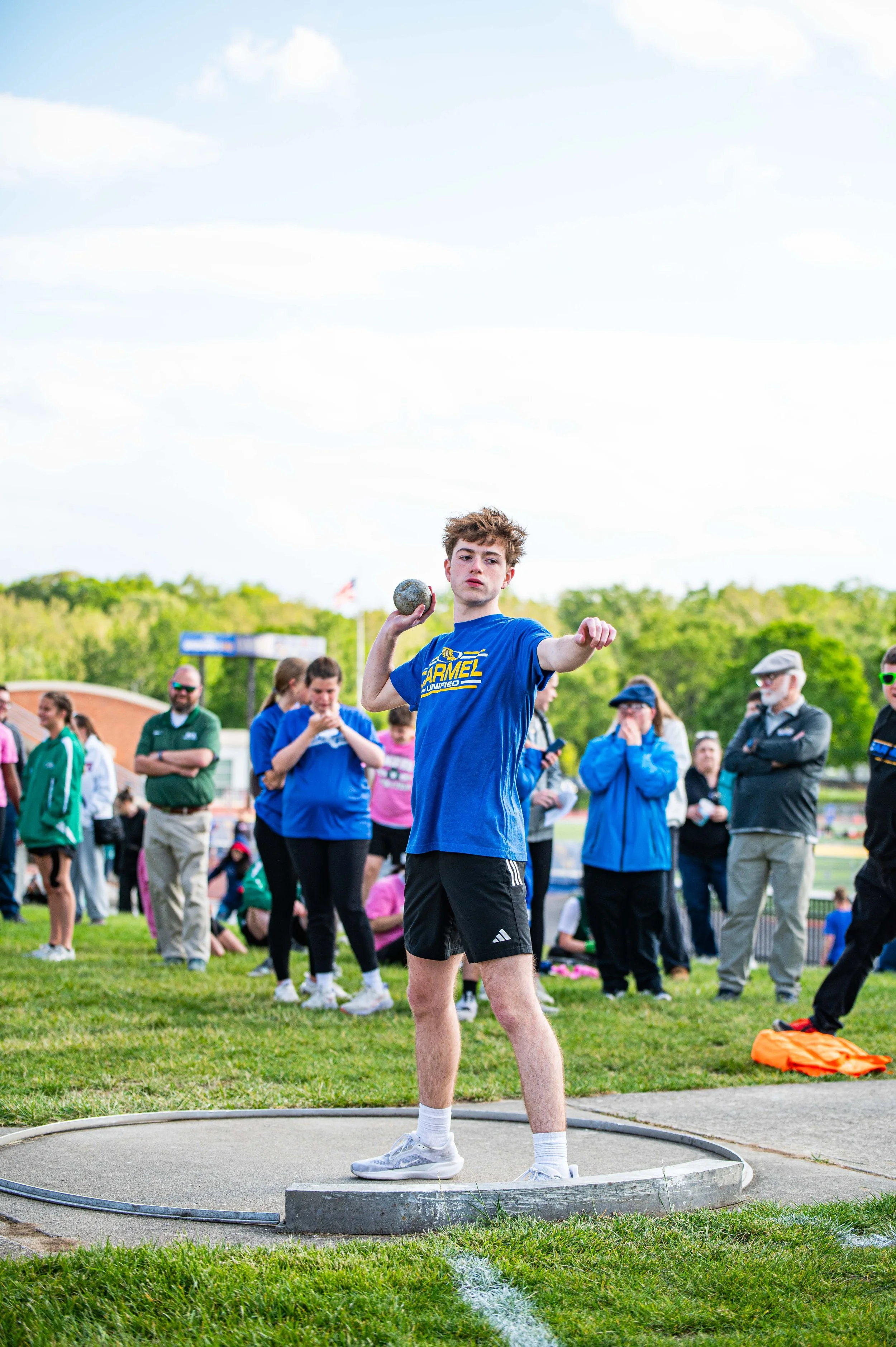 Young male athlete competing in shot put event at track and field meet, surrounded by spectators and fellow athletes, on a grassy field under a partly cloudy sky.