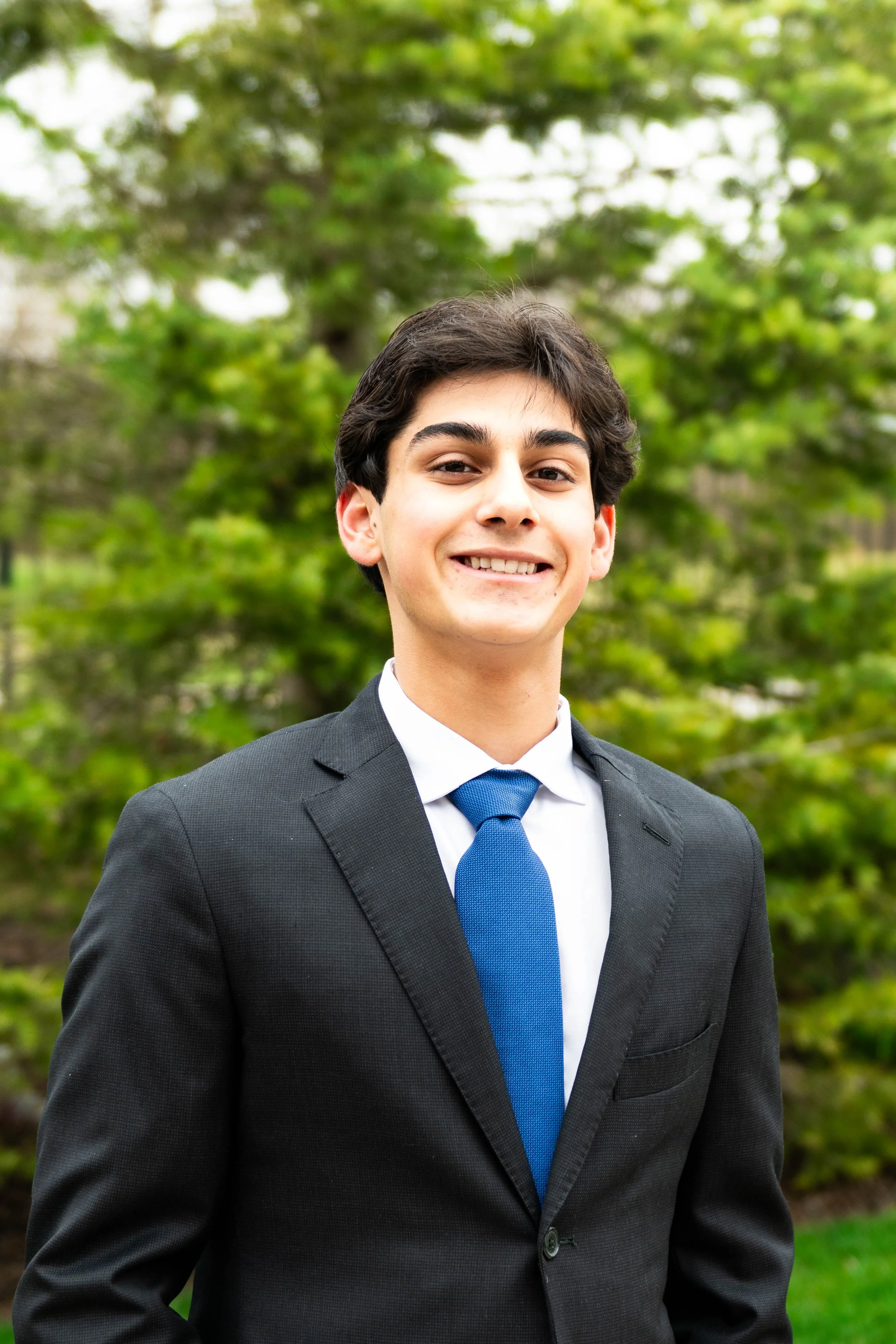 A young man with dark hair and light skin, smiling, dressed in a business suit with a blue tie, standing outdoors with green trees in the background.