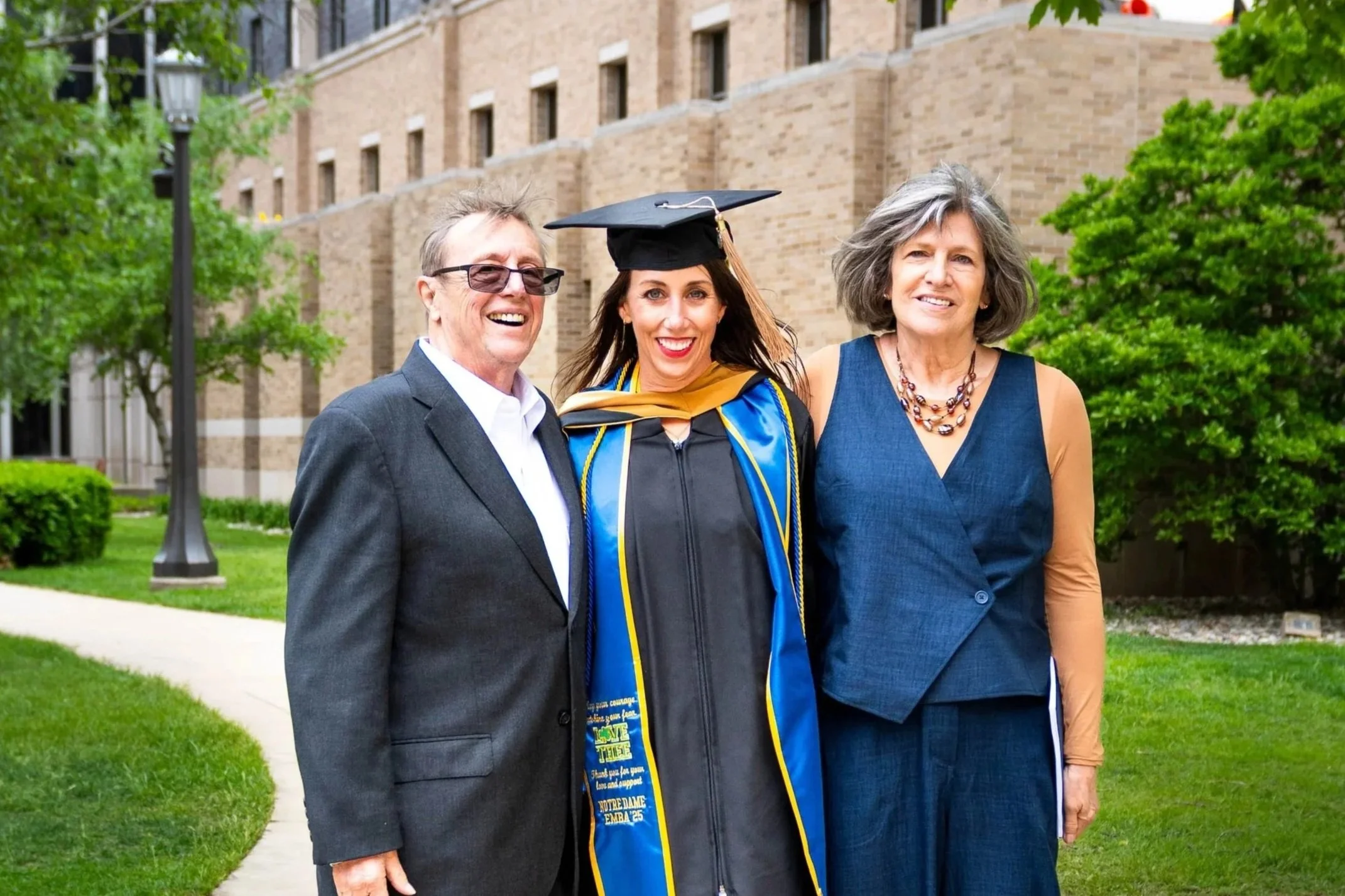 A woman in graduation cap and gown standing between two adults, all smiling and outdoors near a brick building and green trees.
