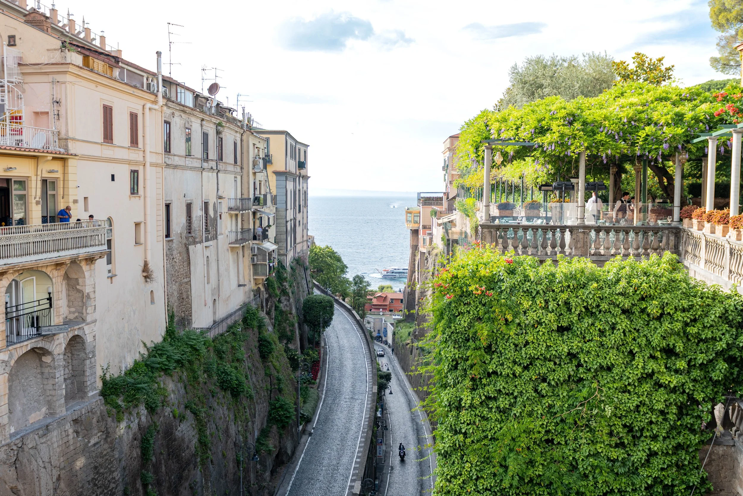 A scenic street view on a hillside overlooking the sea, with historic European-style buildings on the left and lush greenery, including trees and flowering plants, on the right. A cobblestone road runs down the hill with a motorcycle and some cars visible.