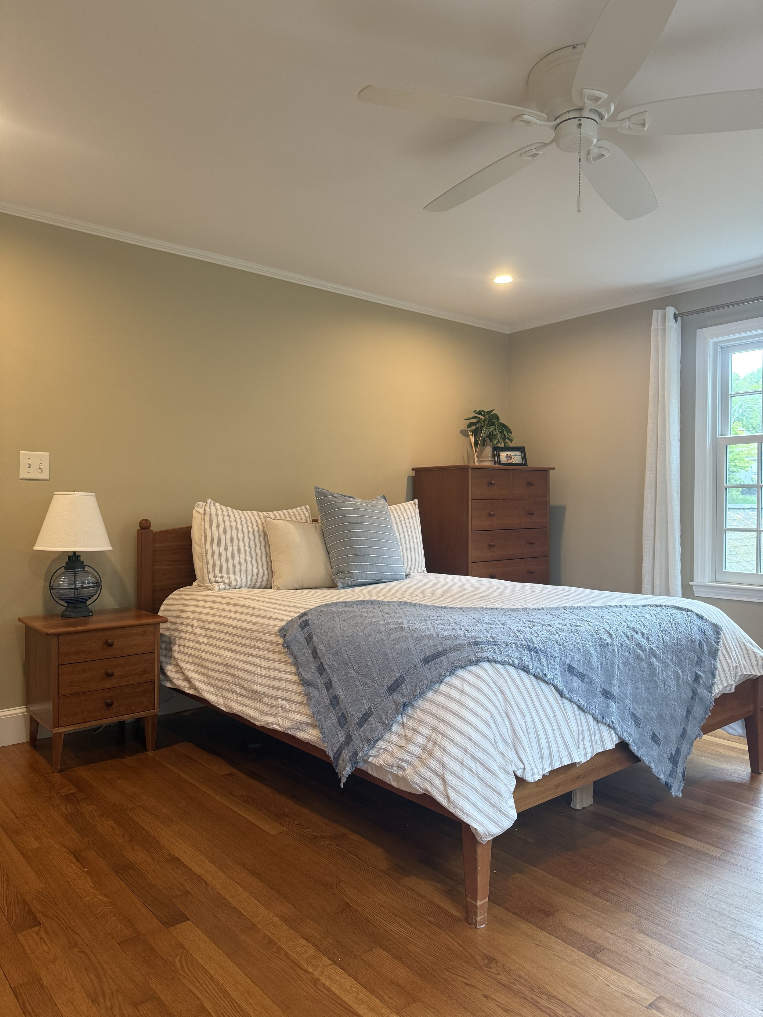 A bedroom with a wooden bed frame, striped bedding, pillows, a nightstand with a lamp, a chest of drawers with a plant and framed photo, a window with white curtains, and a ceiling fan.