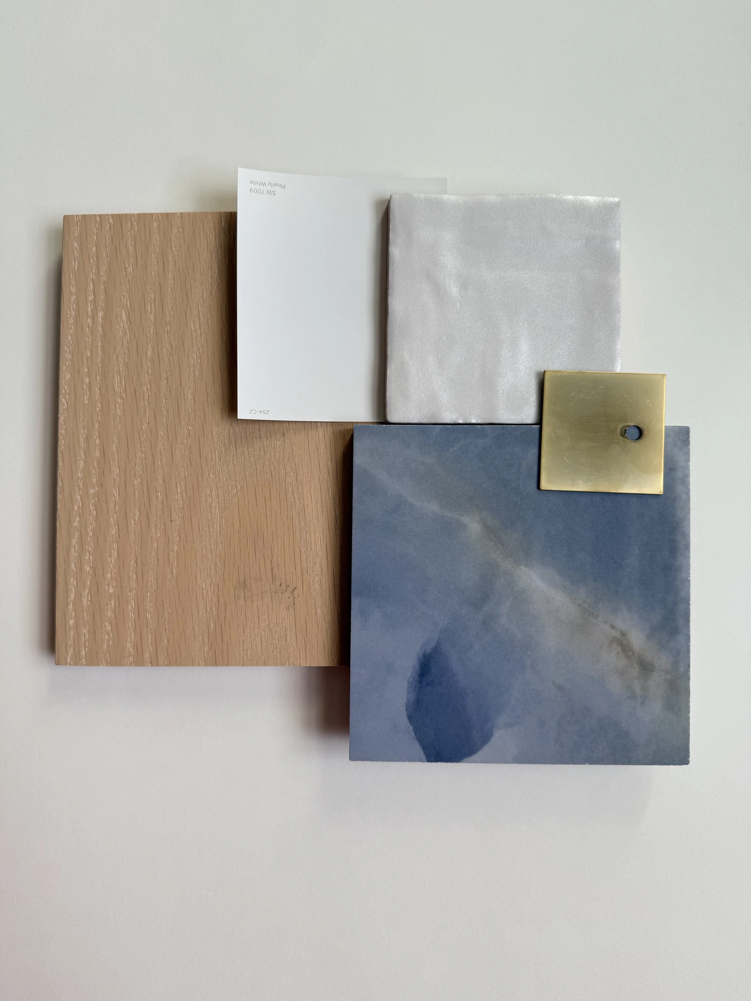 A beautiful flat-lay of various sample materials including a wood-textured large sheet, a white paper, a silver fabric, a gray stone slab, and a brass square with a hole in the center. BY Arlyss in Manchester, Nashua, Salem, and Southern NH.