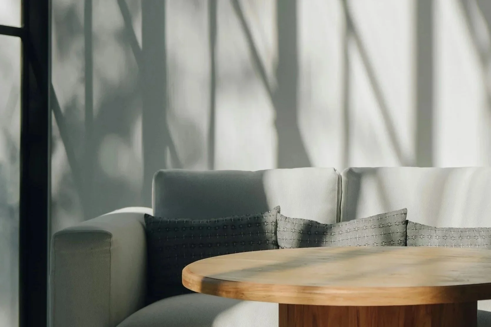 A cozy living room corner with sunlight casting shadows of tree branches on a glass wall, a beige sofa with dark gray cushions, and a wooden round coffee table.