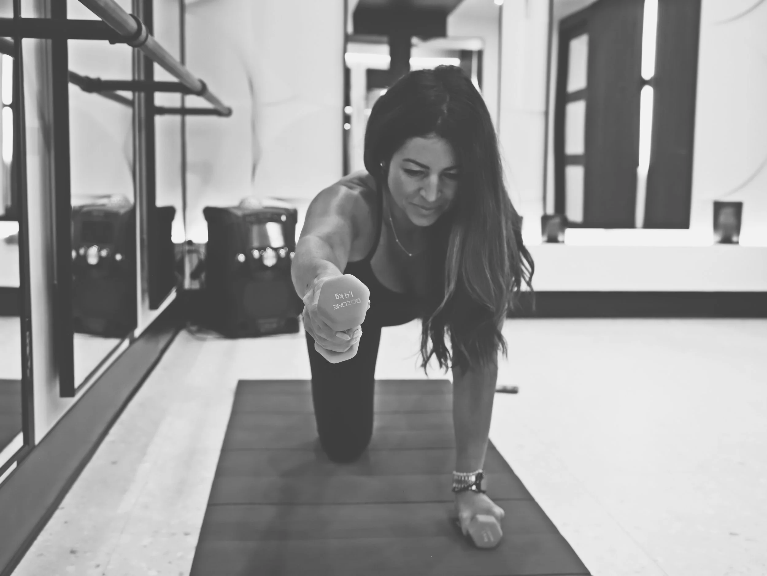 A woman exercising in a gym, kneeling on the floor with her right arm extended forward holding a dumbbell, and her left arm resting on her left knee. The photo is taken at an angle with gym equipment visible in the background.