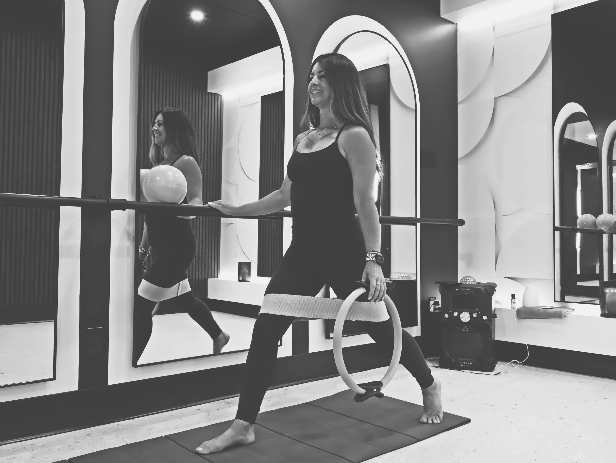 A woman with long hair practicing yoga in front of a mirror. She is standing with one foot on a yoga mat, one knee bent, and her other leg extended straight behind her. She is holding onto a chair with one hand and has a big smile on her face. There are balloons and decorative elements in the background.