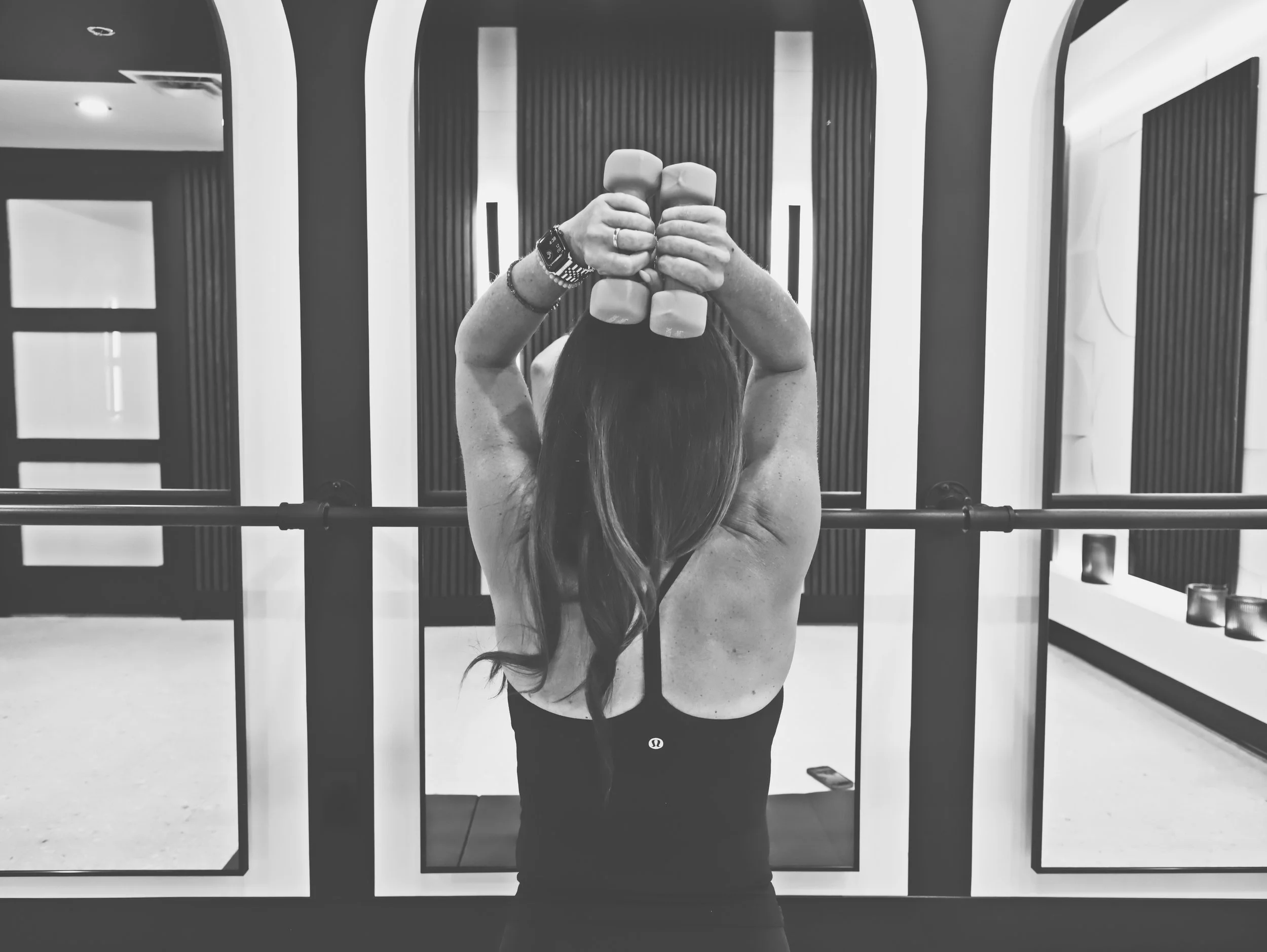 A person taking a mirror selfie in a gym, holding two dumbbells close to their face with both hands. The person is wearing a fitness tracker or smartwatch on their left wrist and rings on their fingers. The background features a modern gym with vertical wall panels and a doorway.