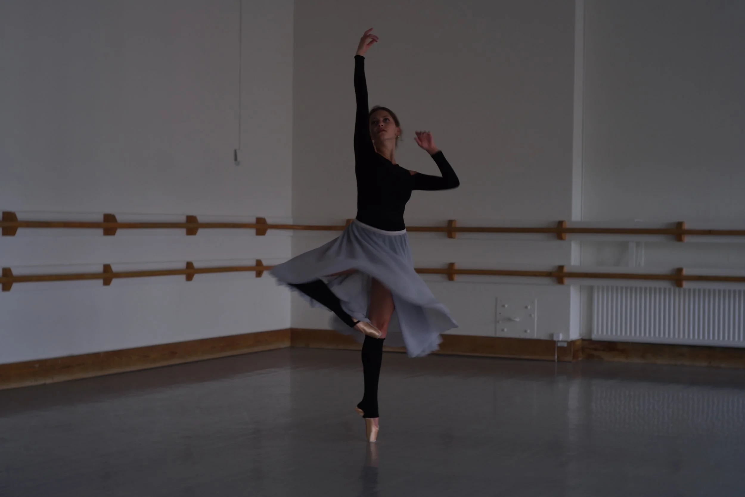 A ballet dancer practicing in a studio with wooden ballet barres on the wall behind her, wearing a black top and a flowing gray skirt, standing en pointe with her left leg supporting her and her right arm raised gracefully.