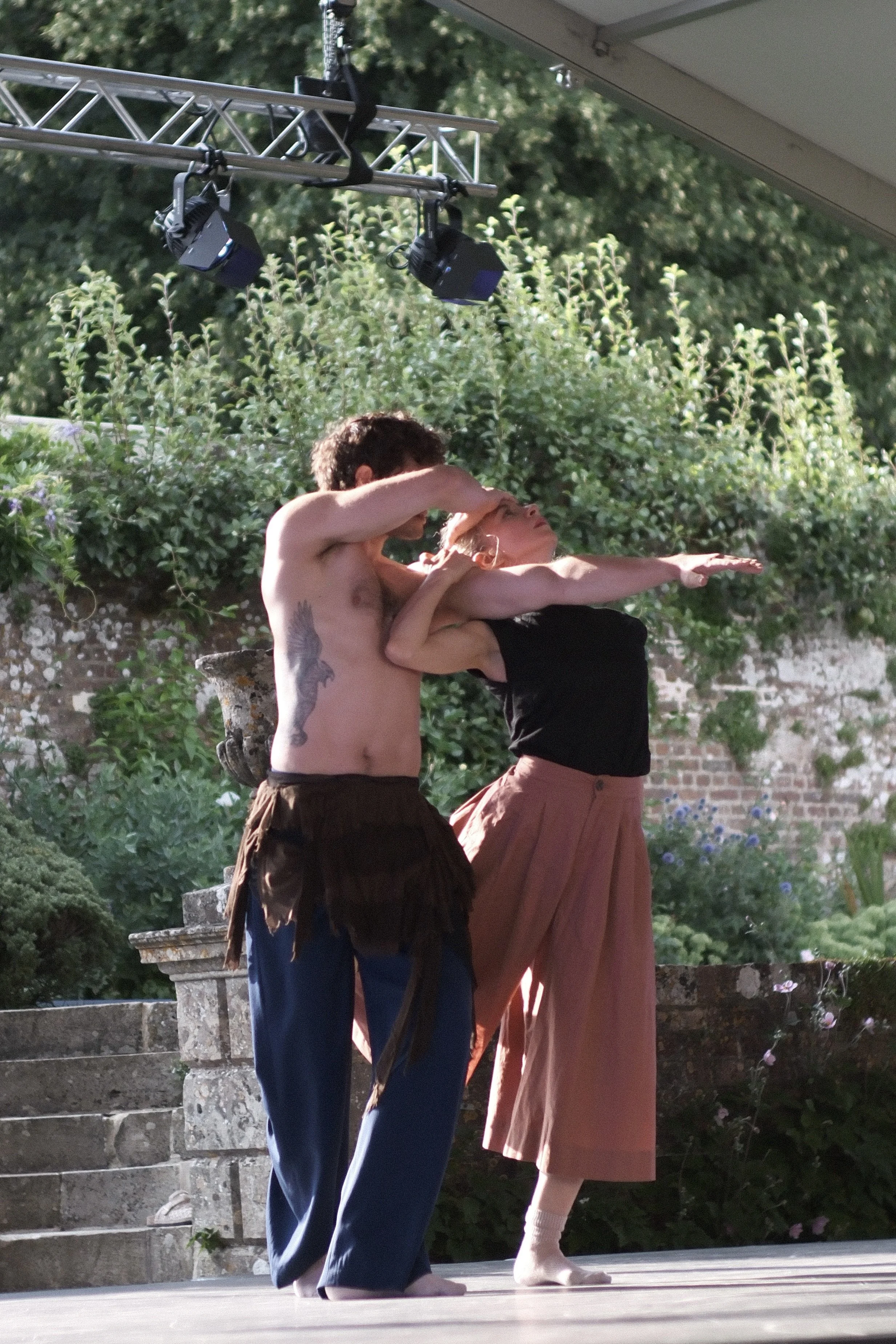 Two dancers, a man and a woman, are performing on a stage outdoors during daylight. The man is shirtless with a tattoo on his side, and the woman is dressed in a black top and a long, flowing peach-colored skirt. They are performing a dance pose with