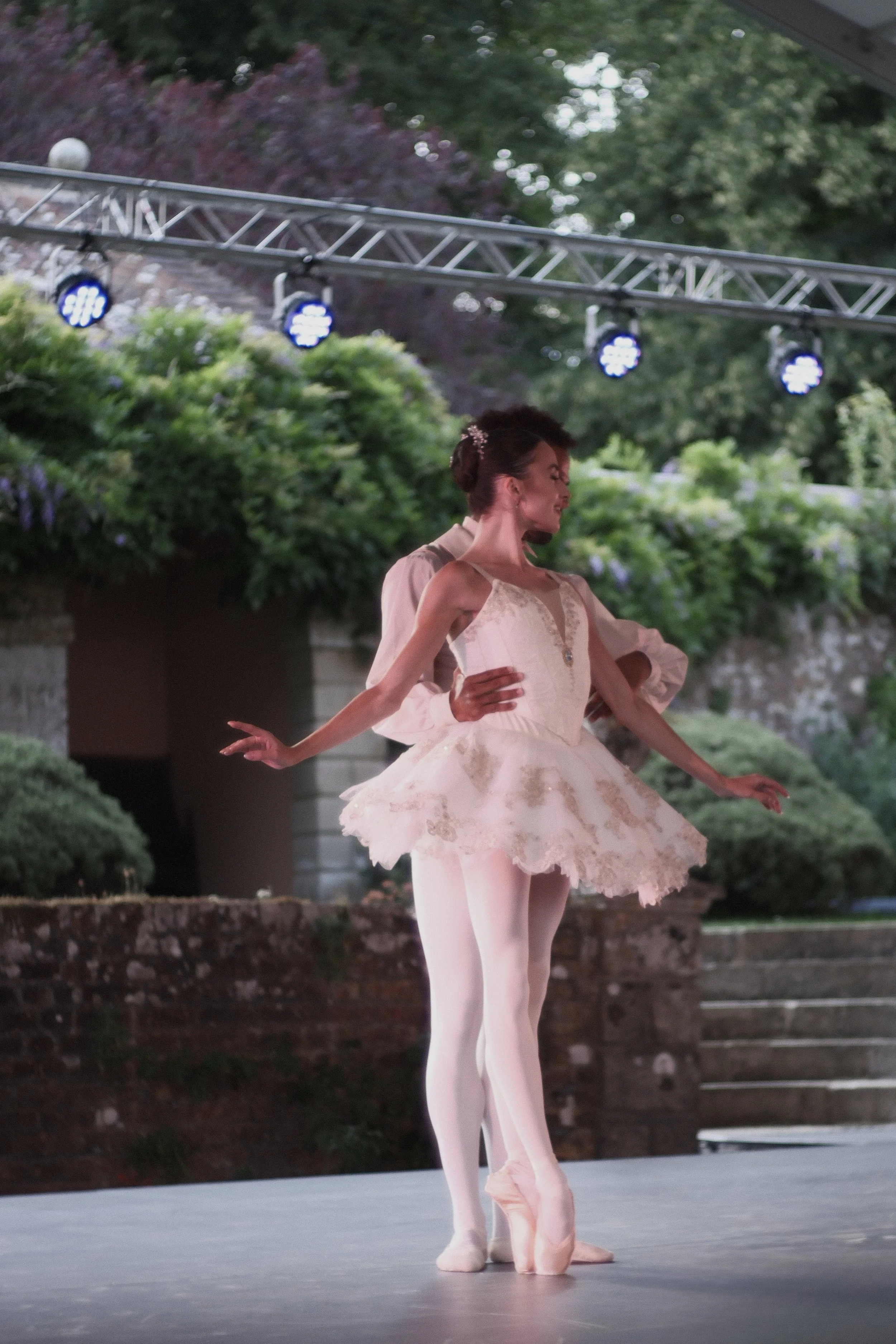 Ballet dancers performing on an outdoor stage at dusk, with trees and stage lighting in the background.