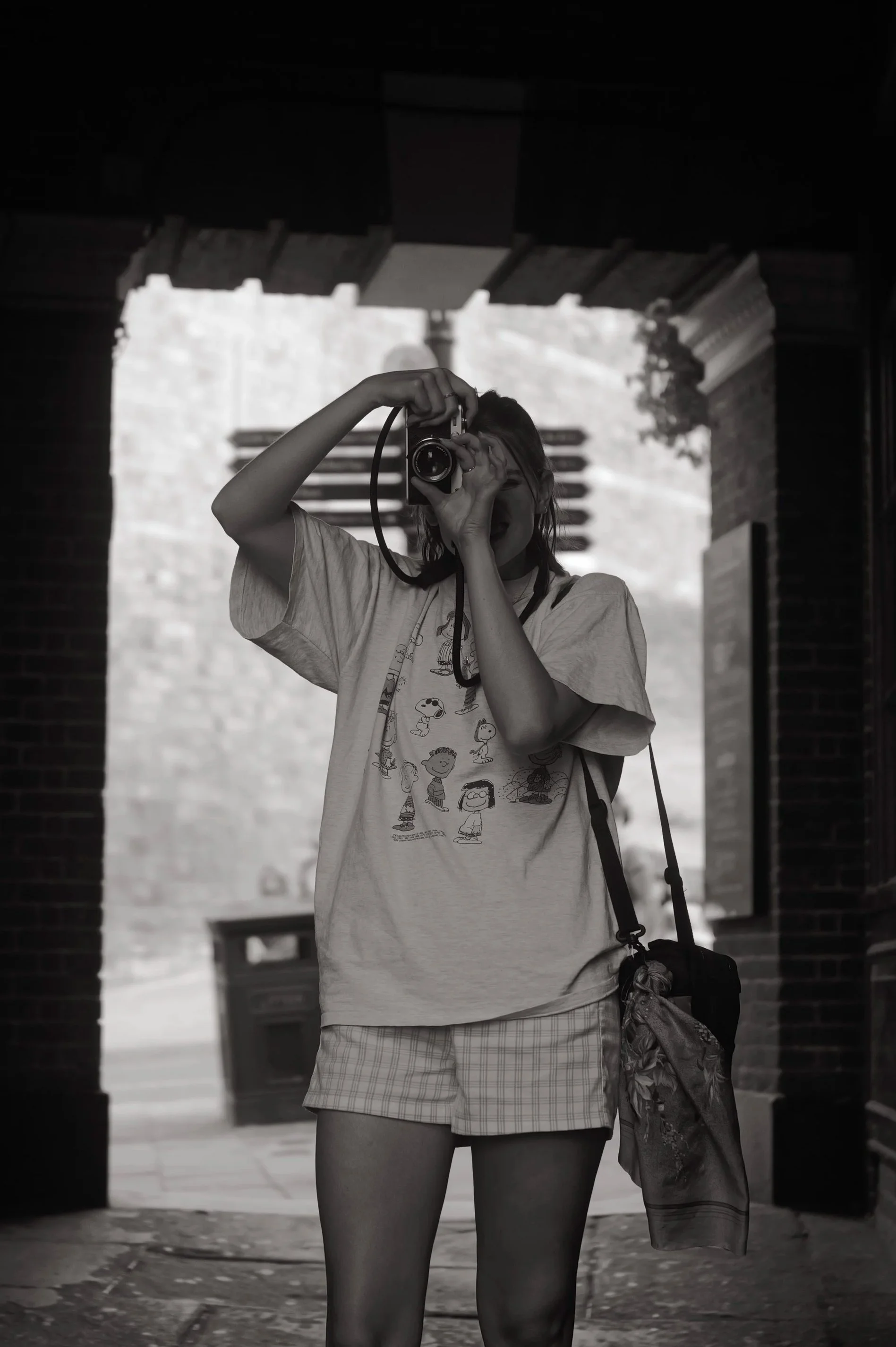 A person taking a selfie with a camera, wearing a graphic t-shirt and plaid shorts, standing in an outdoor area with brick structures and a bench in the background.