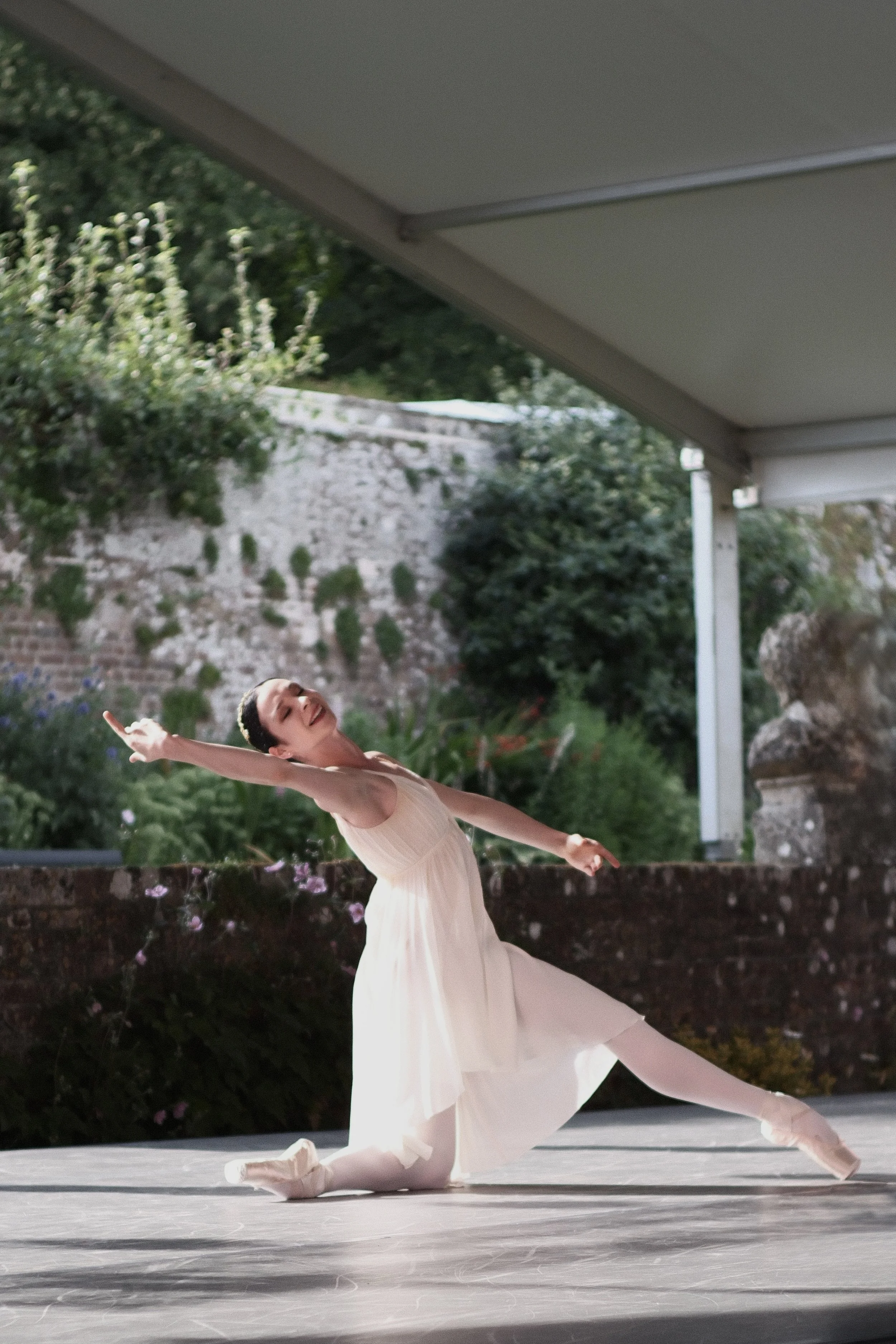 A young female ballet dancer performing a dance pose outdoors on a stage, wearing a light-colored dress and ballet shoes, with her arm extended and one leg bent behind her.