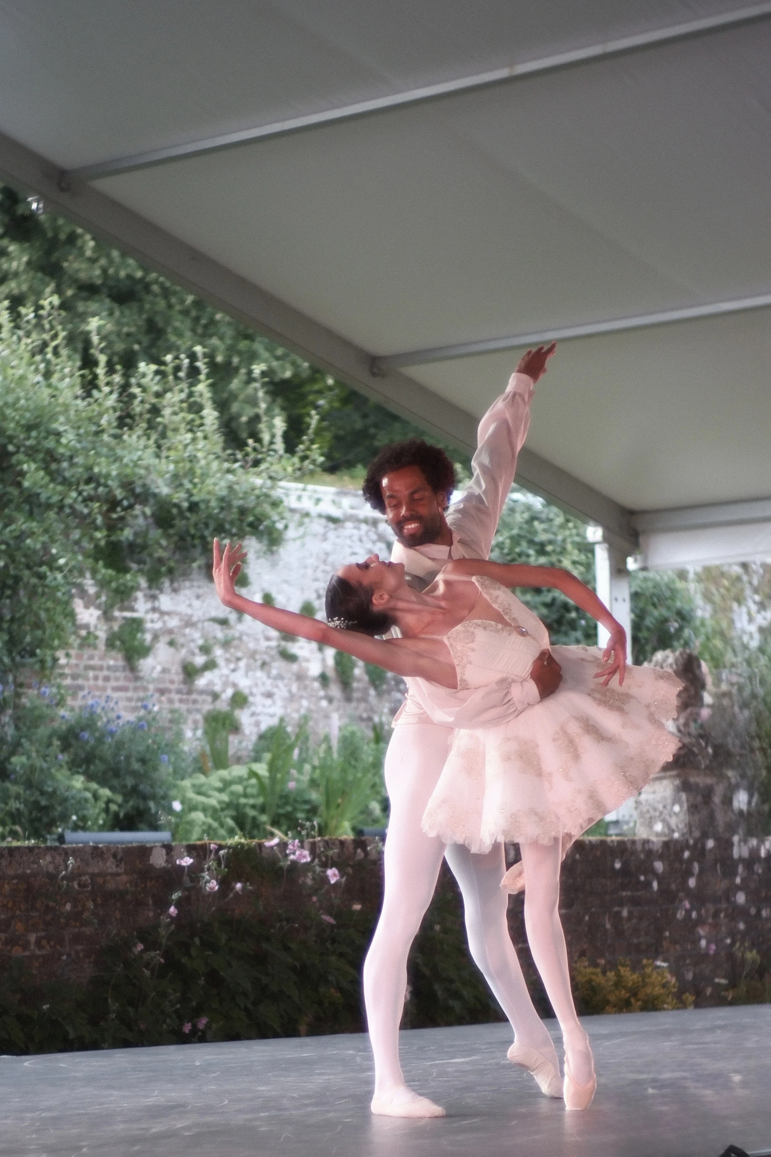 Ballet dancers performing on stage outdoors, with a male dancer lifting a female dancer, both in ballet poses, surrounded by greenery and flowers.