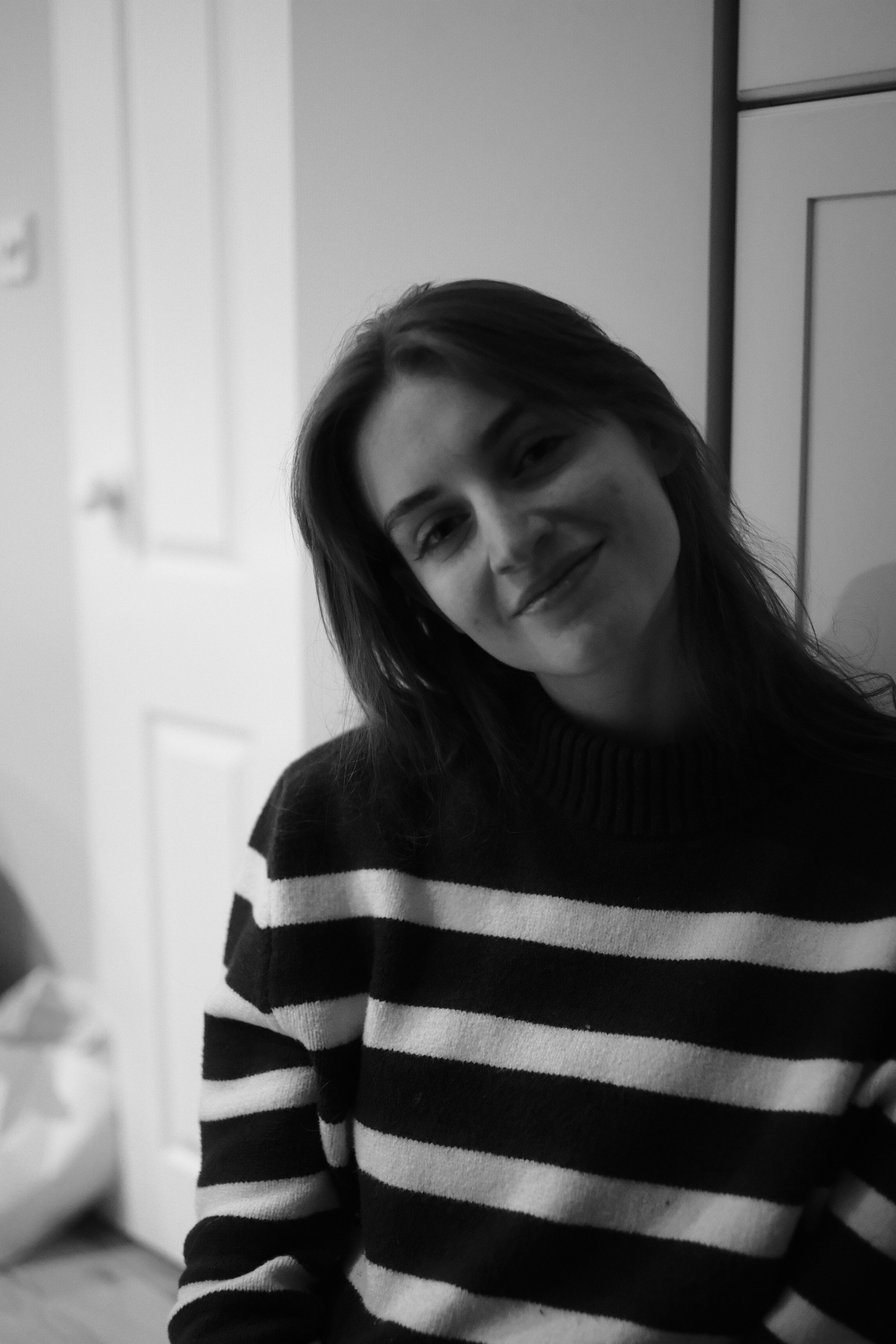 Black and white photo of a smiling woman with long hair, wearing a striped sweater, sitting indoors.