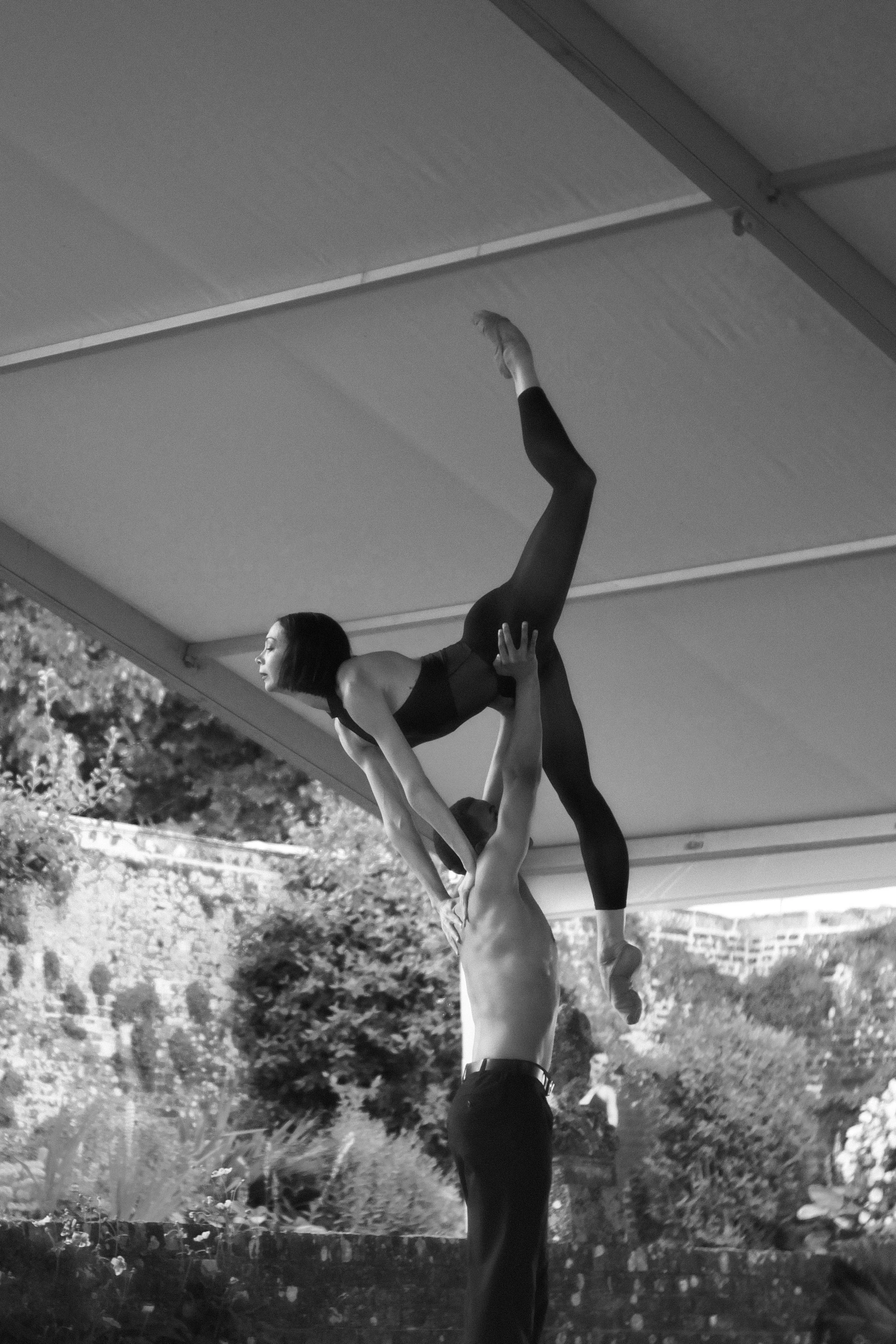 A woman performing an acro yoga pose with a man outdoors under a pavilion, with trees and grass in the background, captured in black and white.