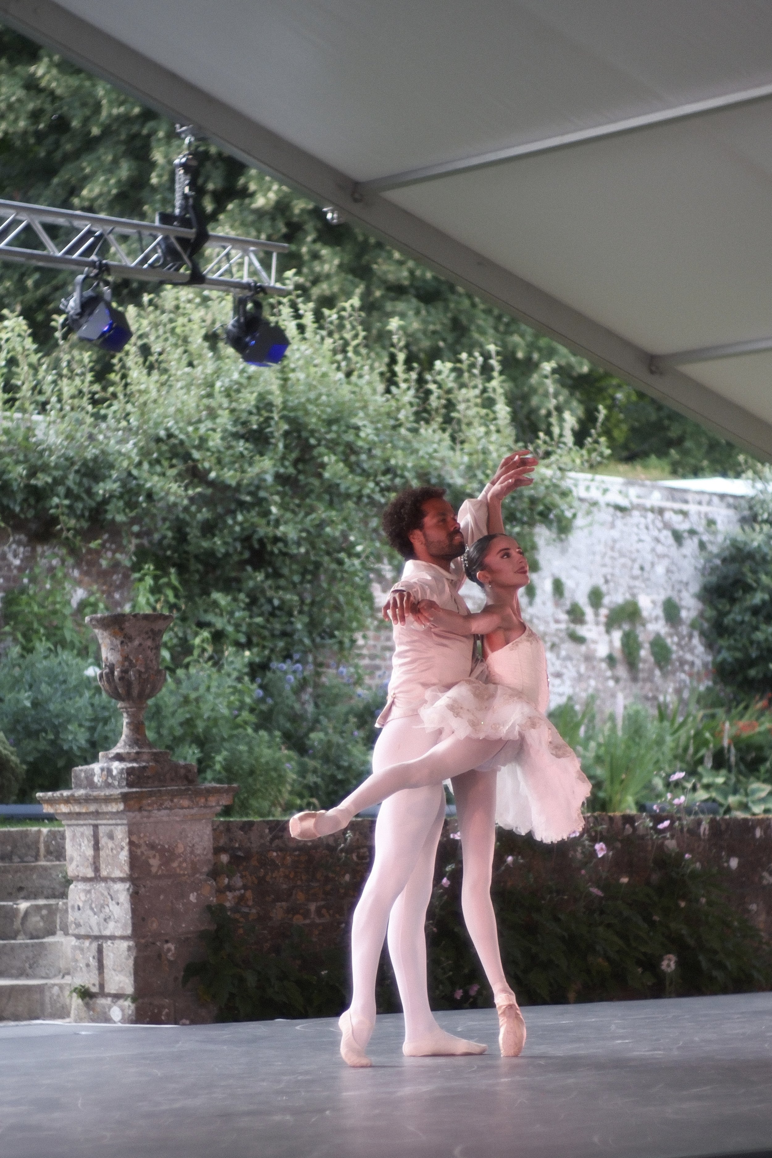 Two ballet dancers perform outdoors on a stage, with one dancer lifting the other. The dancers wear light-colored costumes and tights. The setting features greenery, a stone pedestal with a decorative urn, and stage lighting equipment overhead.