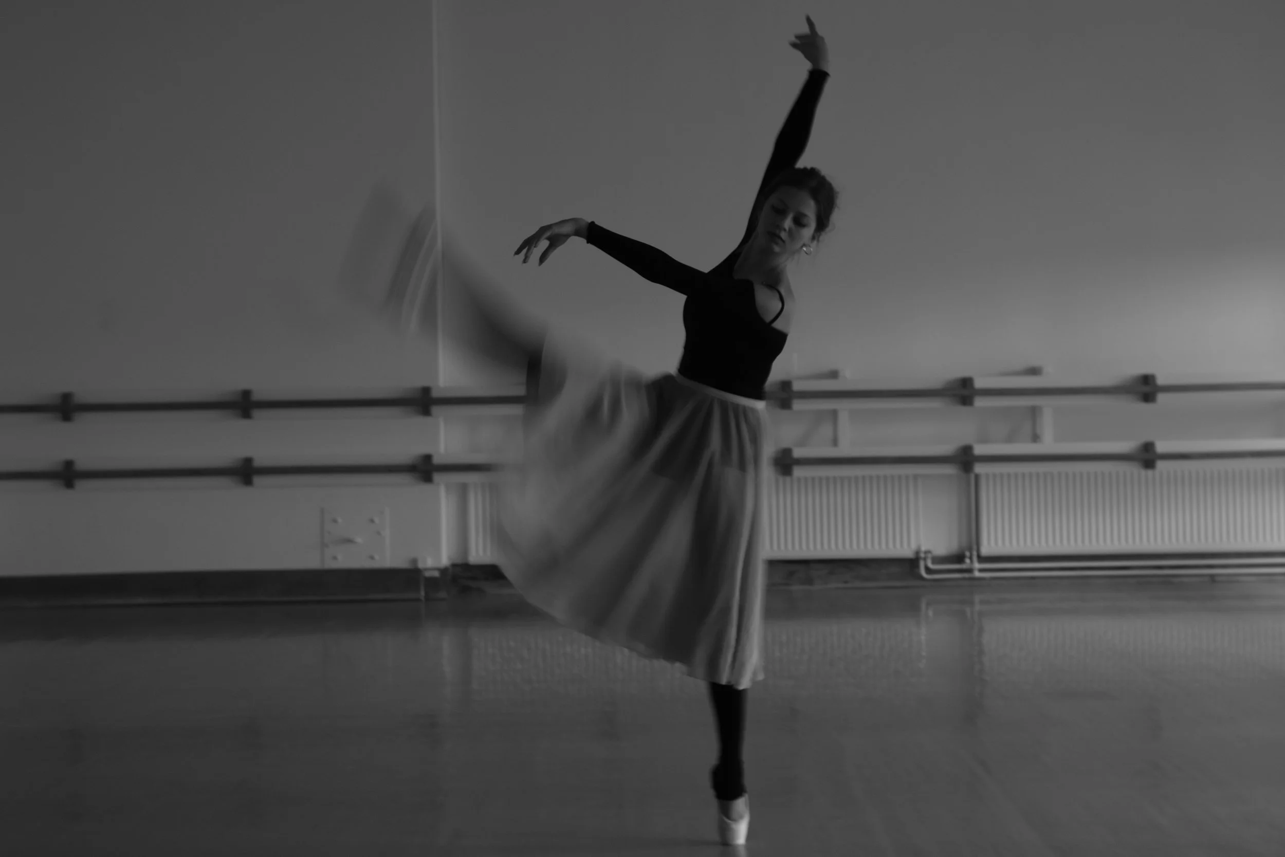 A ballerina practicing a move at a dance studio, with her arm raised gracefully and her leg extended, wearing a black top and a flowy skirt, with ballet bars on the wall behind her.